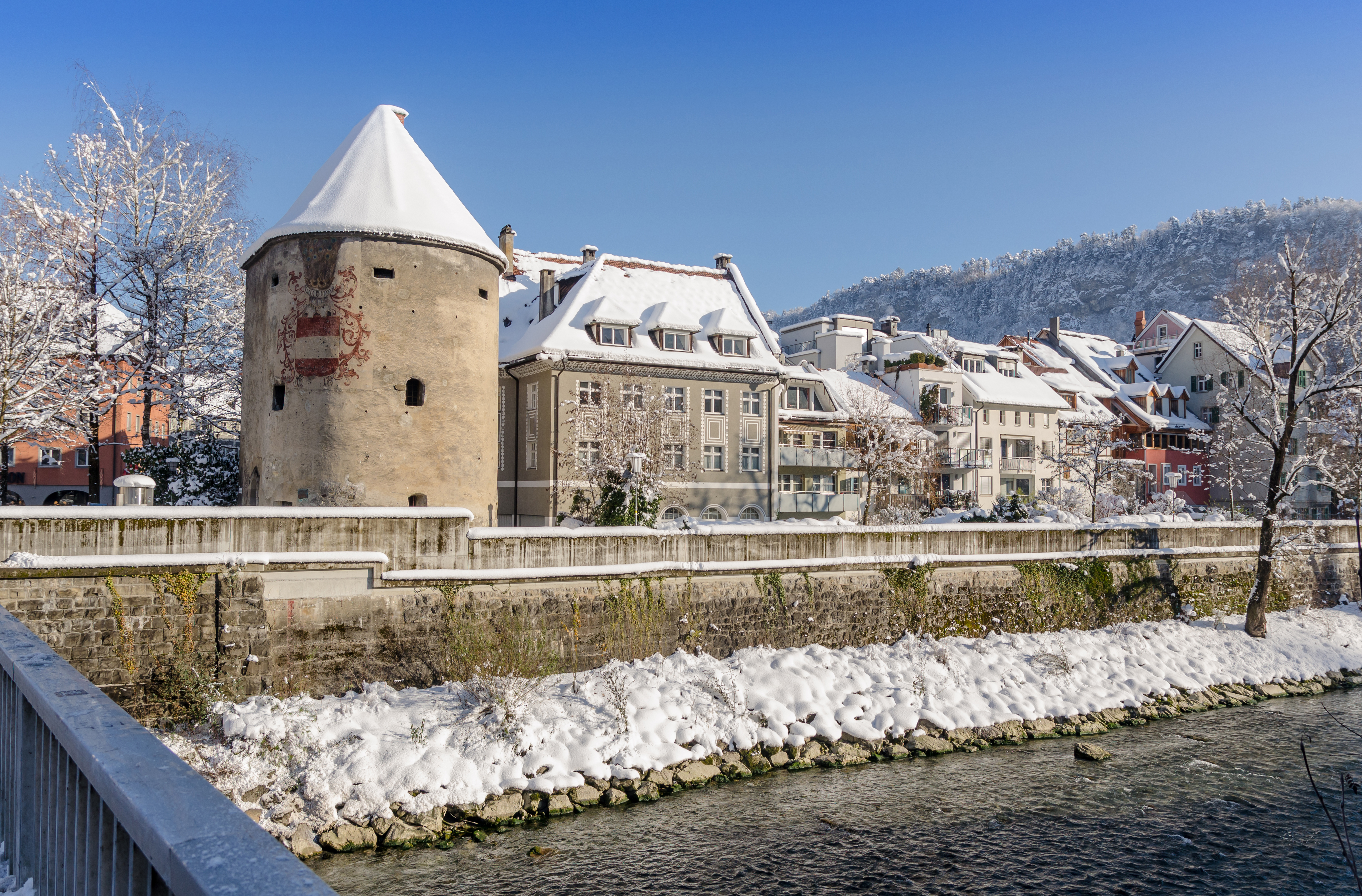 Snedækket historisk bykerne i Feldkirch, Vorarlberg med middelalderligt tårn og flodpromenade omgivet af Alperne om vinteren