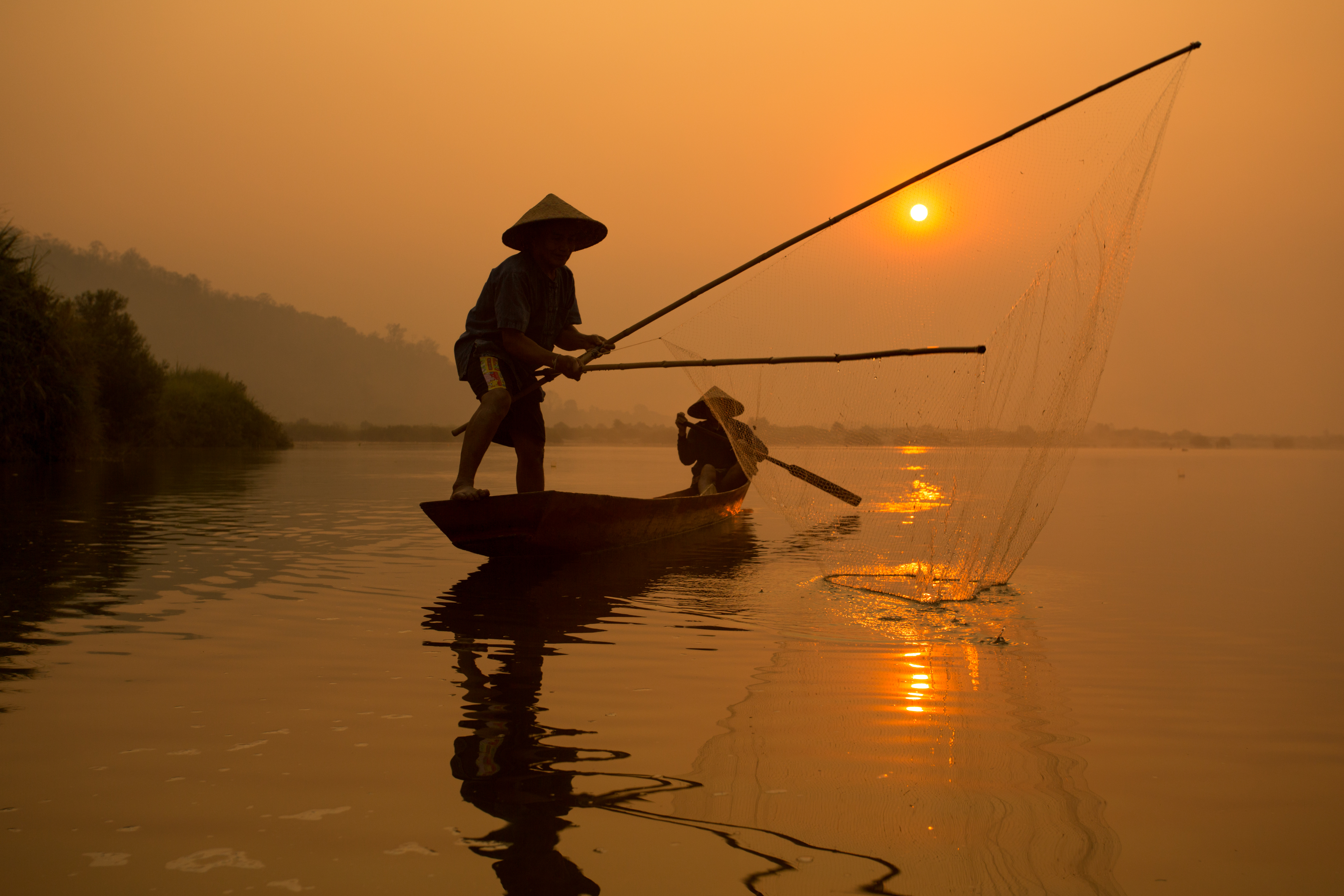 To fiskere i traditionel træbåd kaster net ved solnedgang på flod i Thailand