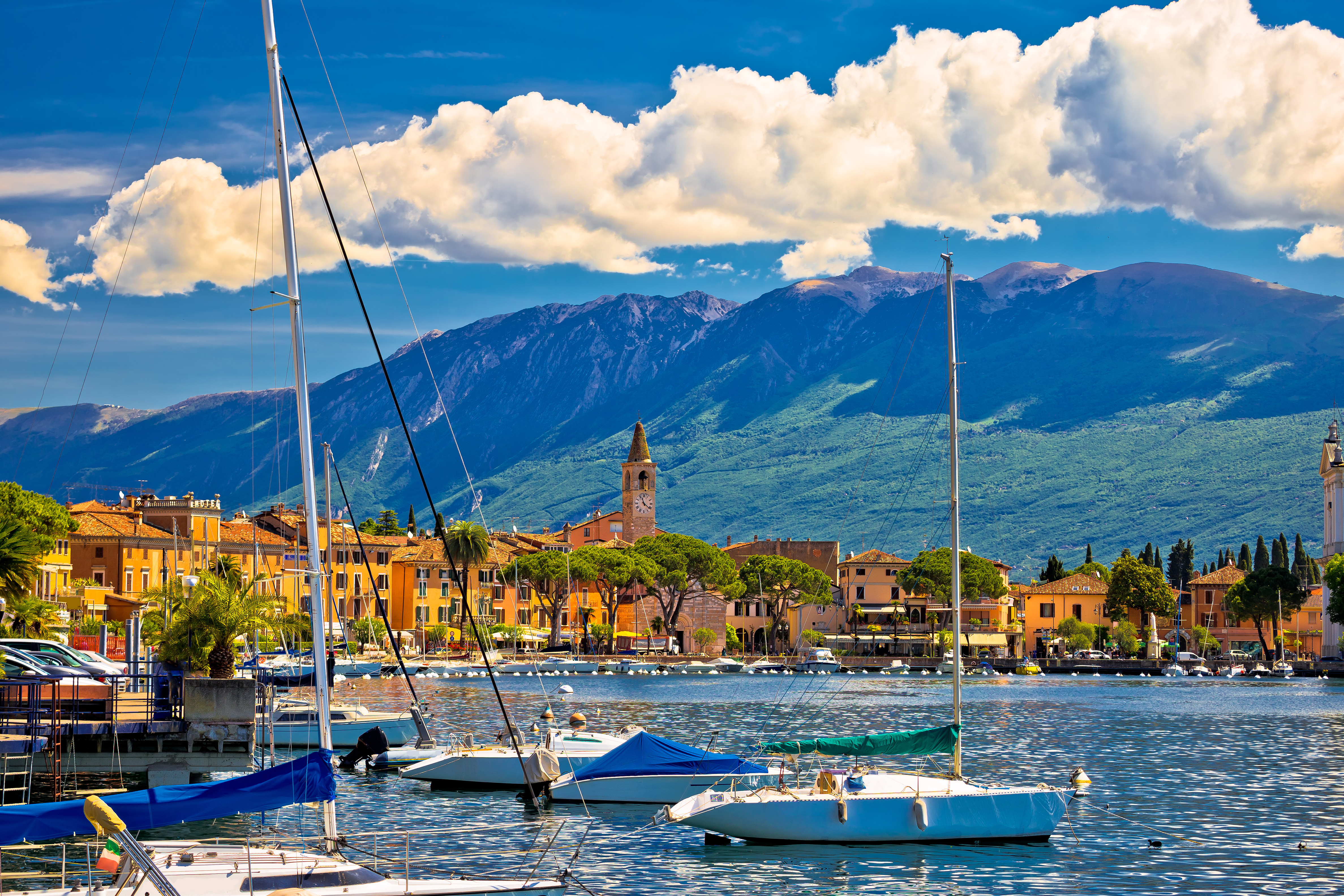 Malerisk havn ved Gardasøen i Italien med sejlbåde, historiske bygninger og storslåede bjerglandskaber i Lombardiet på en solrig sommerdag