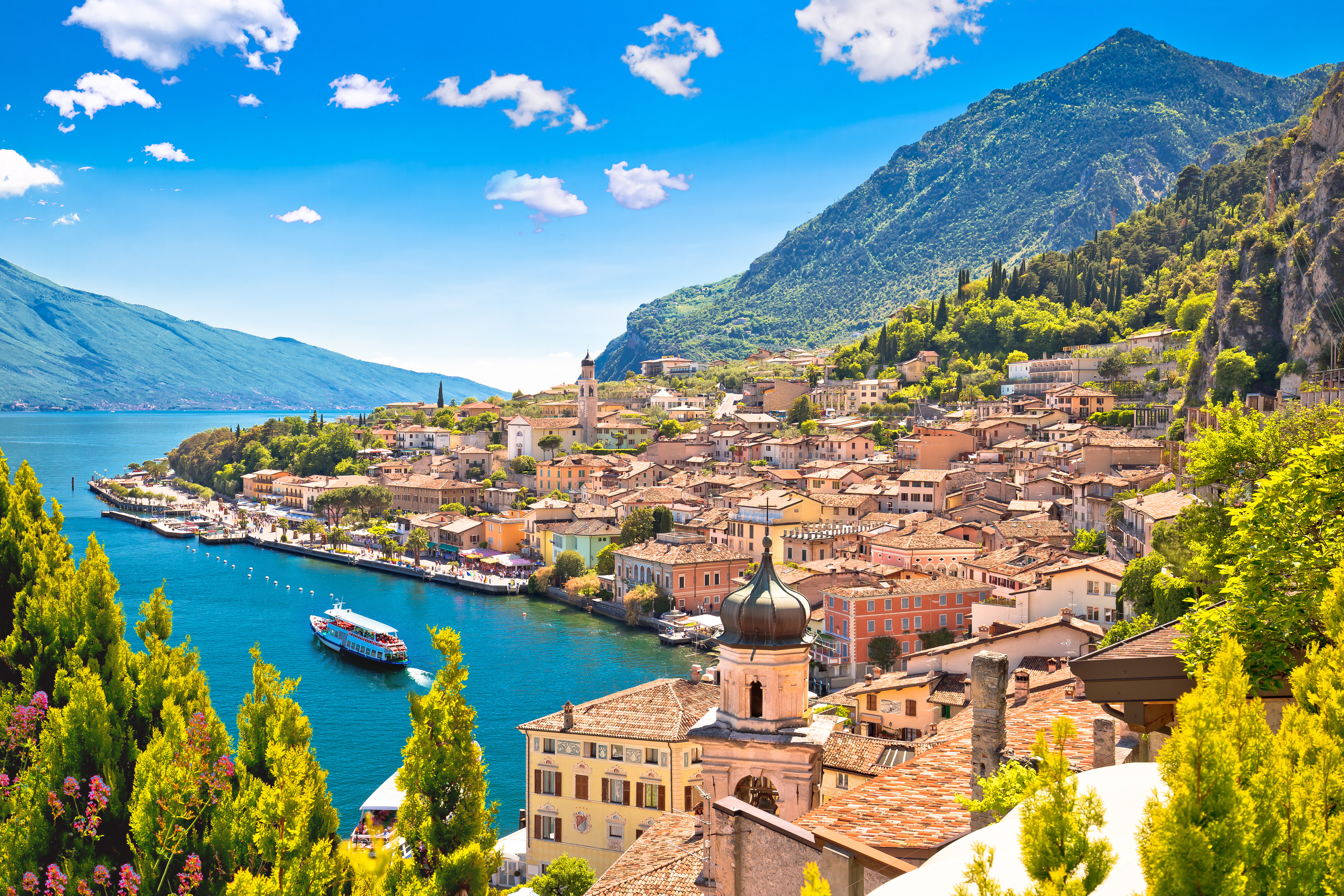Malerisk udsigt over Limone sul Garda med farverige huse, kirkespir og en lystbåd på det turkisblå Gardasø med bjerge i baggrunden