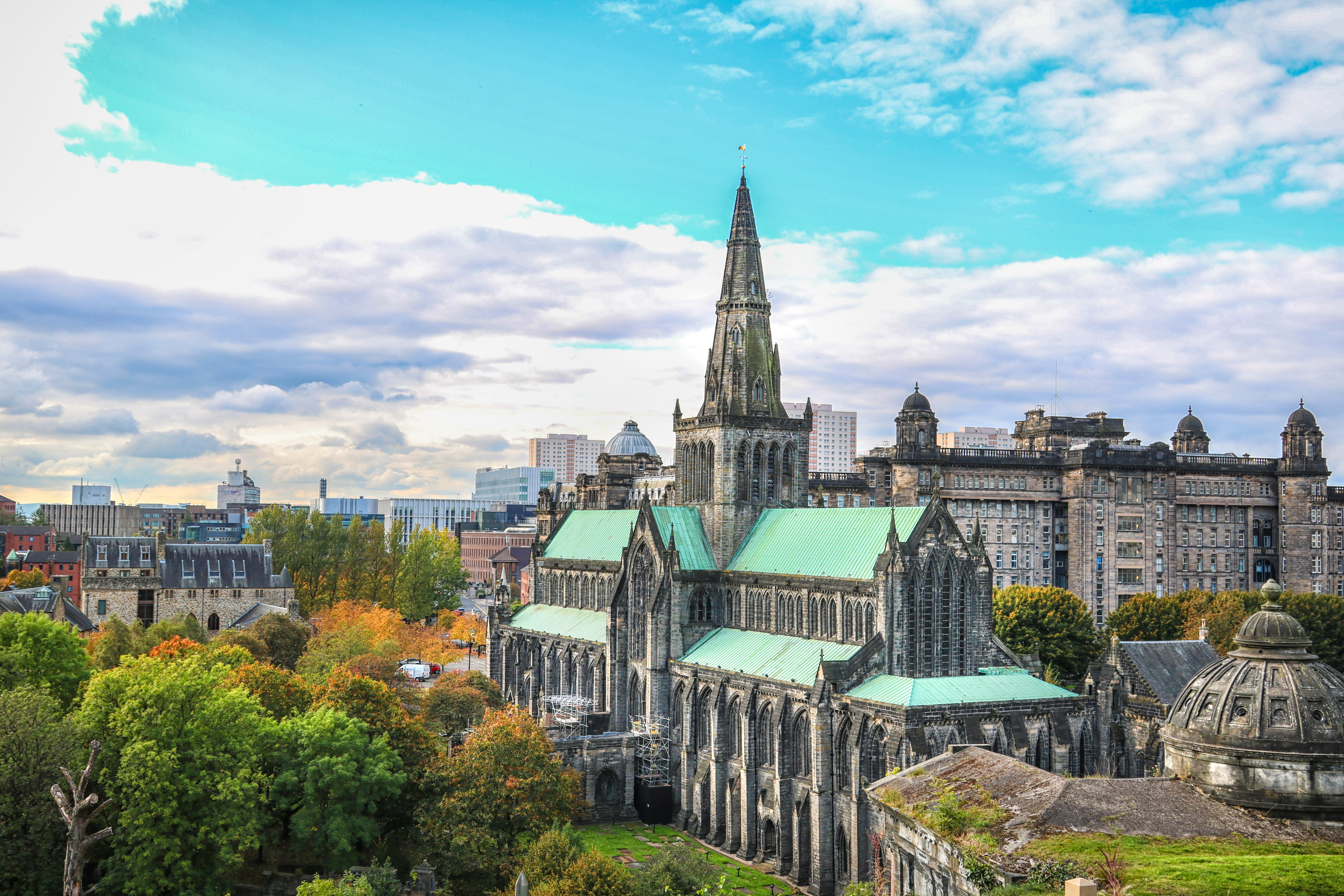 Smukt overblik over Glasgow Cathedral med gotisk spir og grønne kobbertage omgivet af efterårsfarver og Glasgow skyline i Skotland
