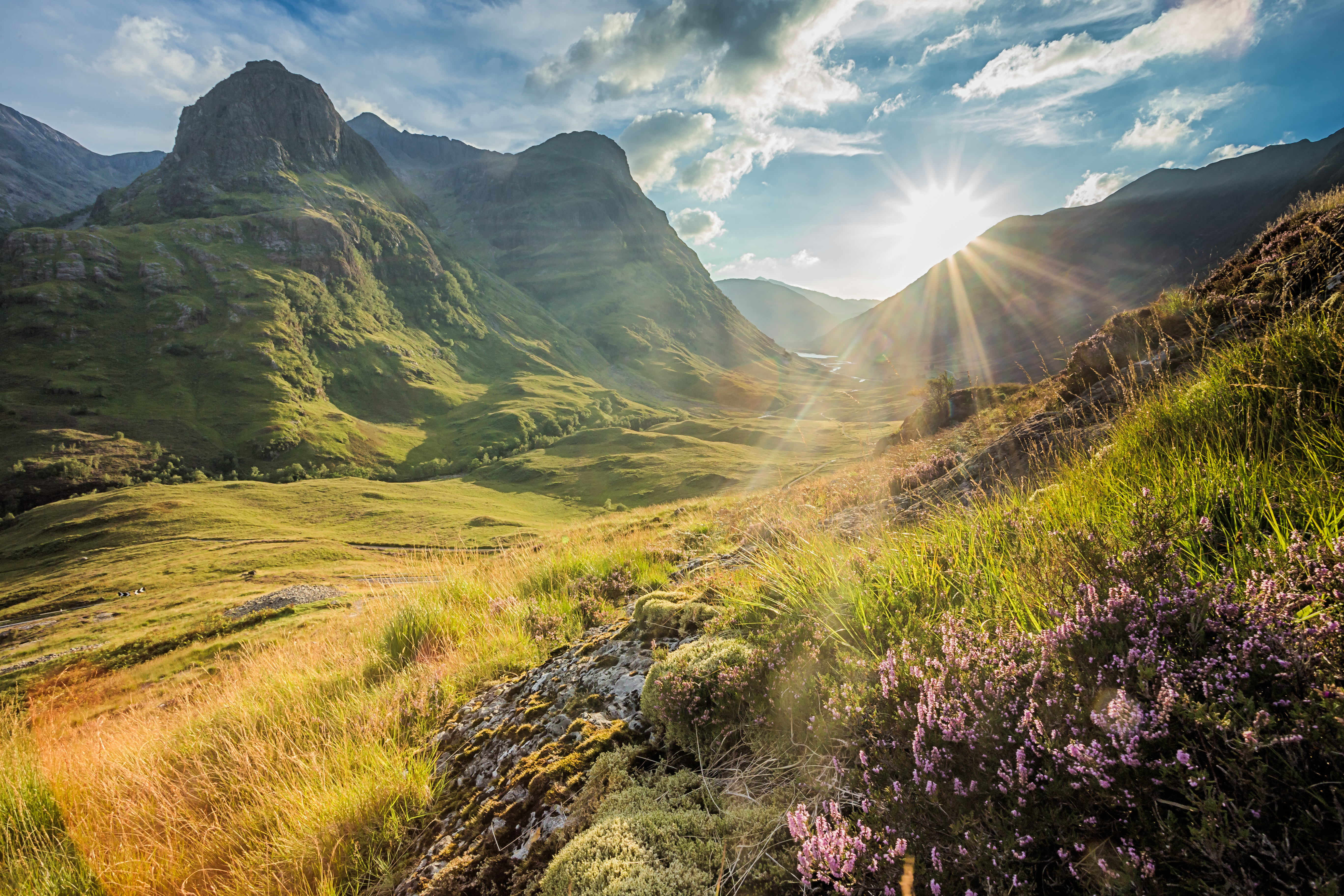Imponerende udsigt over den grønne Glencoe-dal i det skotske højland, omgivet af de markante Three Sisters bjerge og med en snoet vej der fører gennem det naturskønne landskab