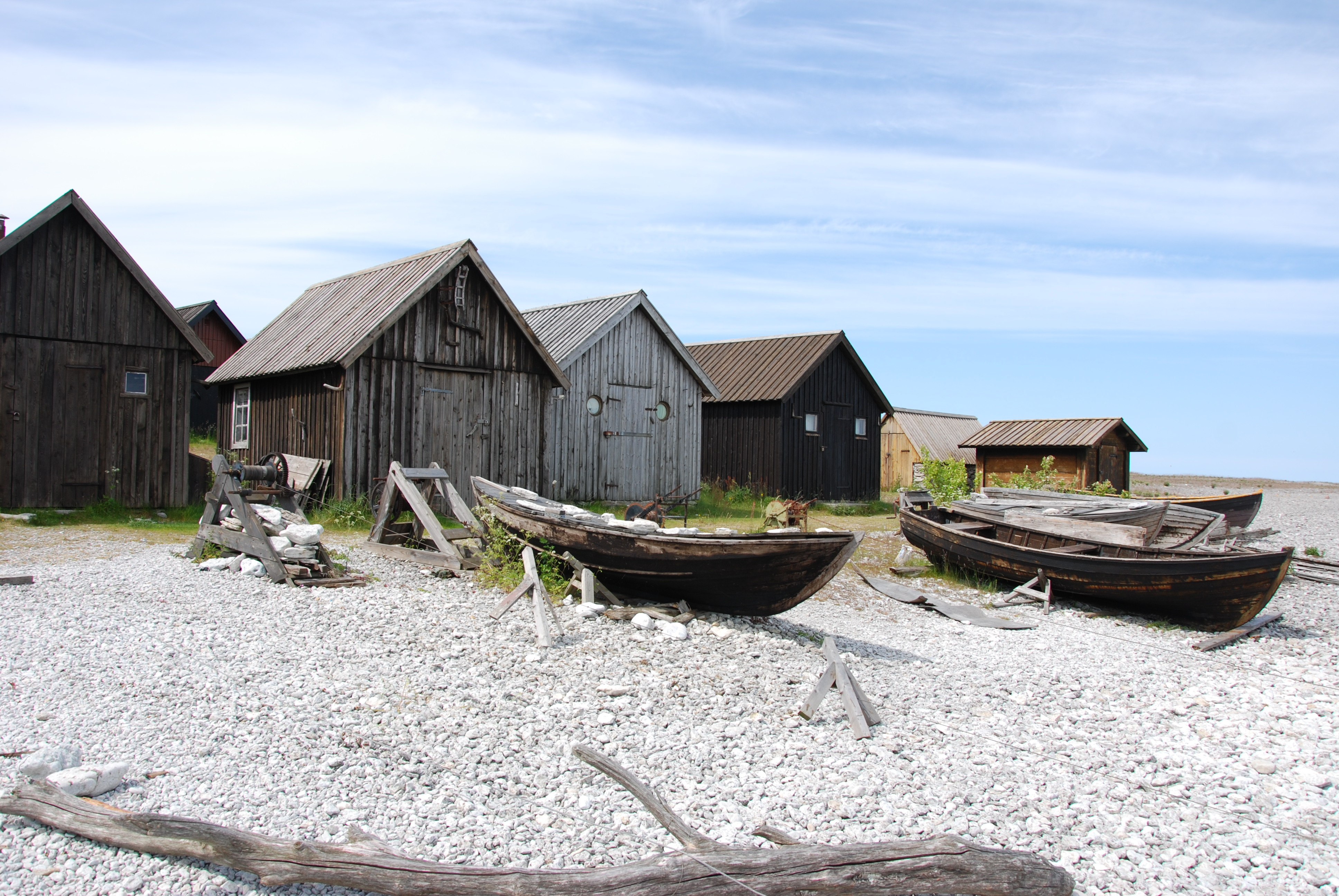 Historiske træfiskerbåde på pebblestrand foran vejrbidte fiskerihuse ved Helgumannen, Gotland, Sverige