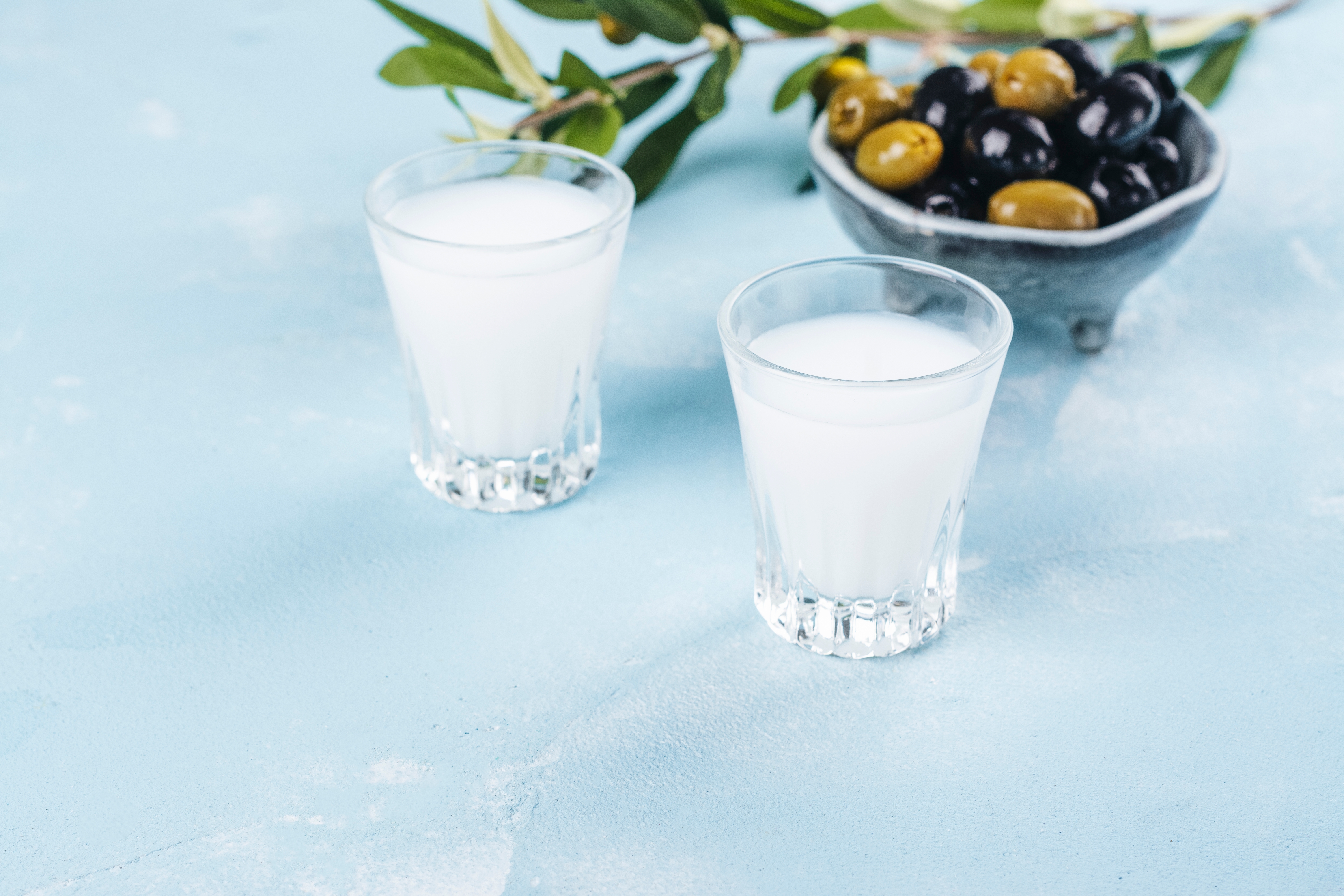 Klassisk græsk ouzo serveret i shotglas med en skål blandede oliven og olivenkviste på blå baggrund
