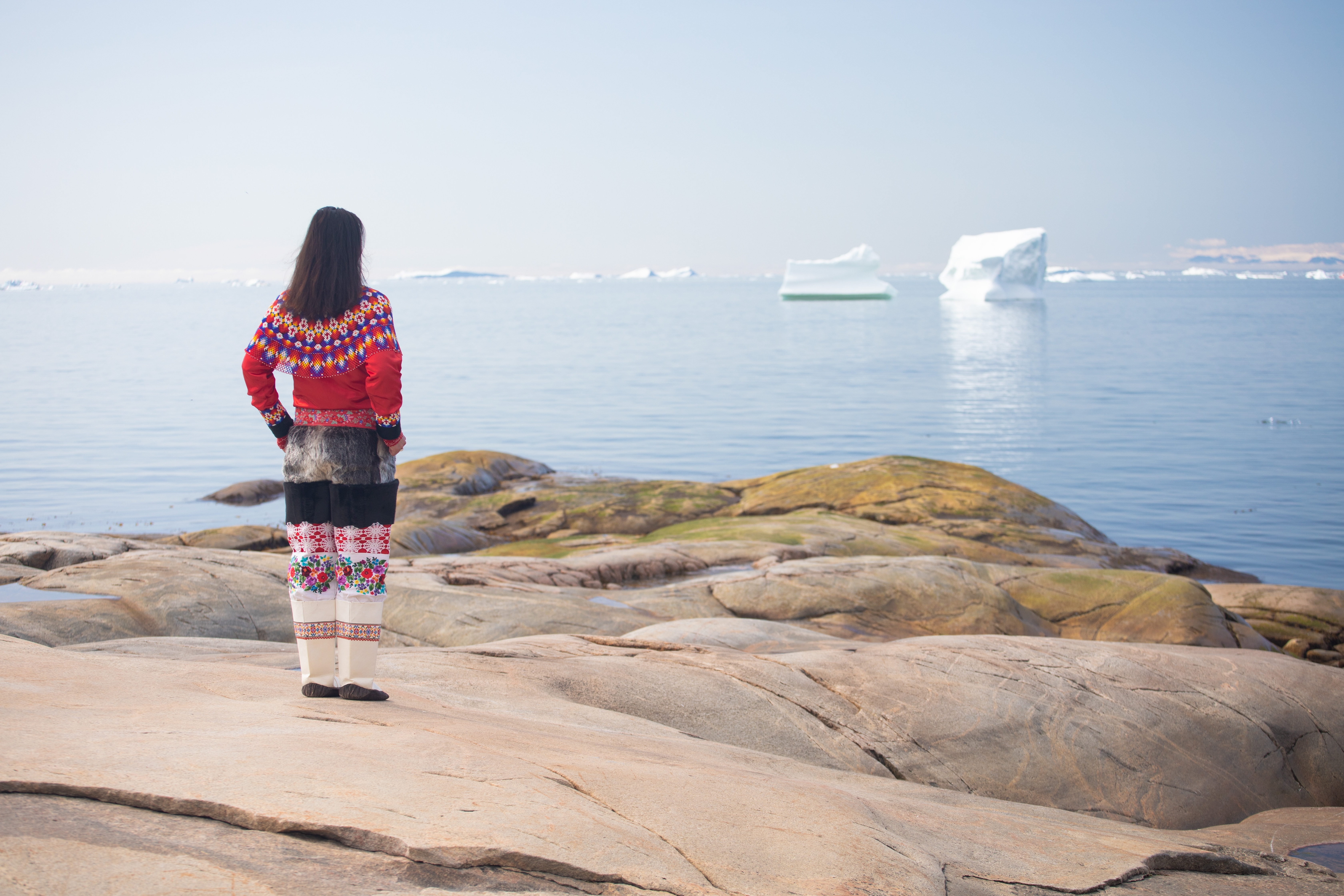 Ung kvinde i traditionel grønlandsk dragt står på klipperne ved Ilulissat og kigger ud over isfjorden med flydende isbjerge