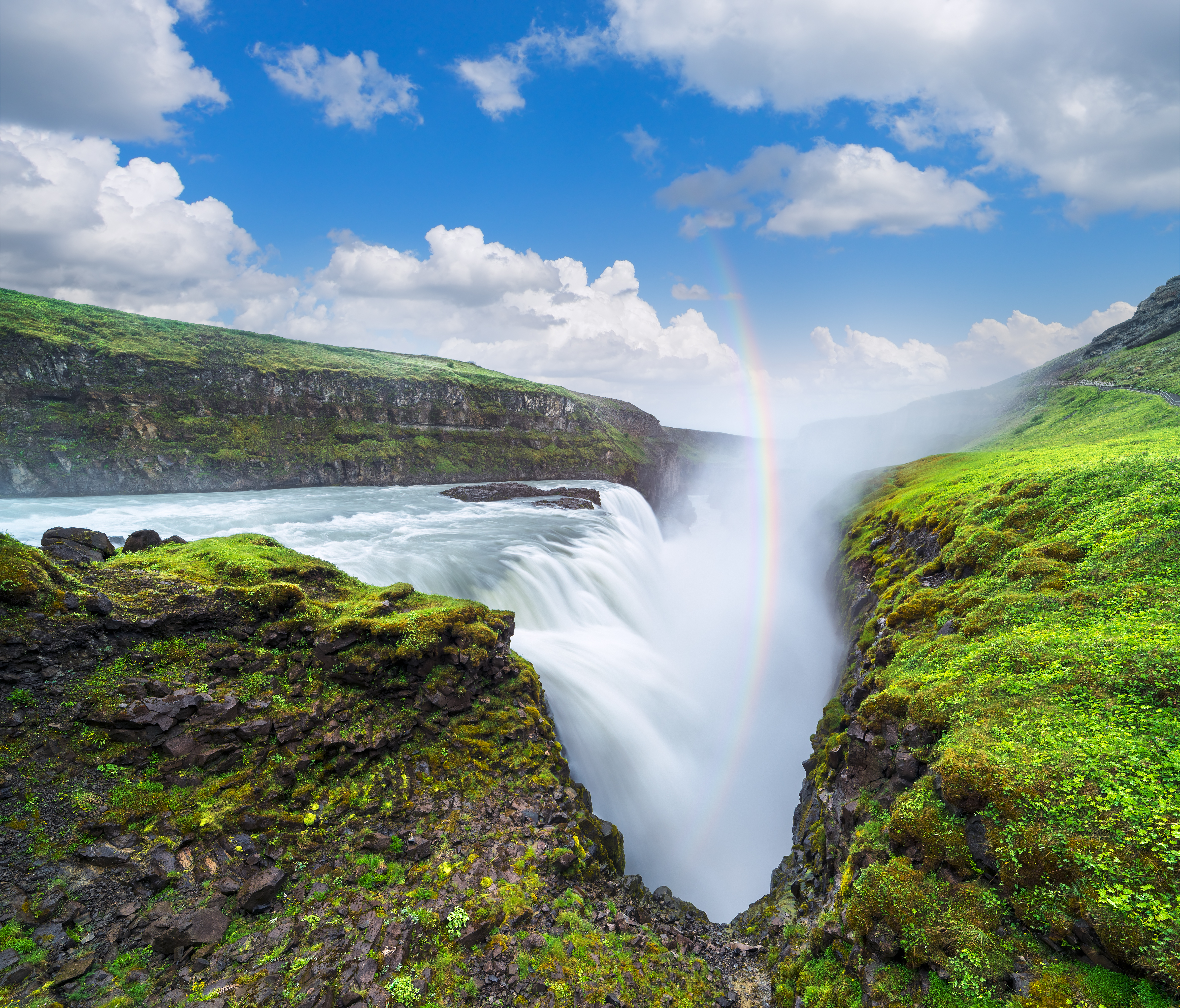 Udsigt over det imponerende Gullfoss vandfald på Island, hvor vandet styrter ned i en dyb kløft med en regnbue i baggrunden