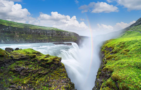 Udsigt over det imponerende Gullfoss vandfald på Island, hvor vandet styrter ned i en dyb kløft med en regnbue i baggrunden
