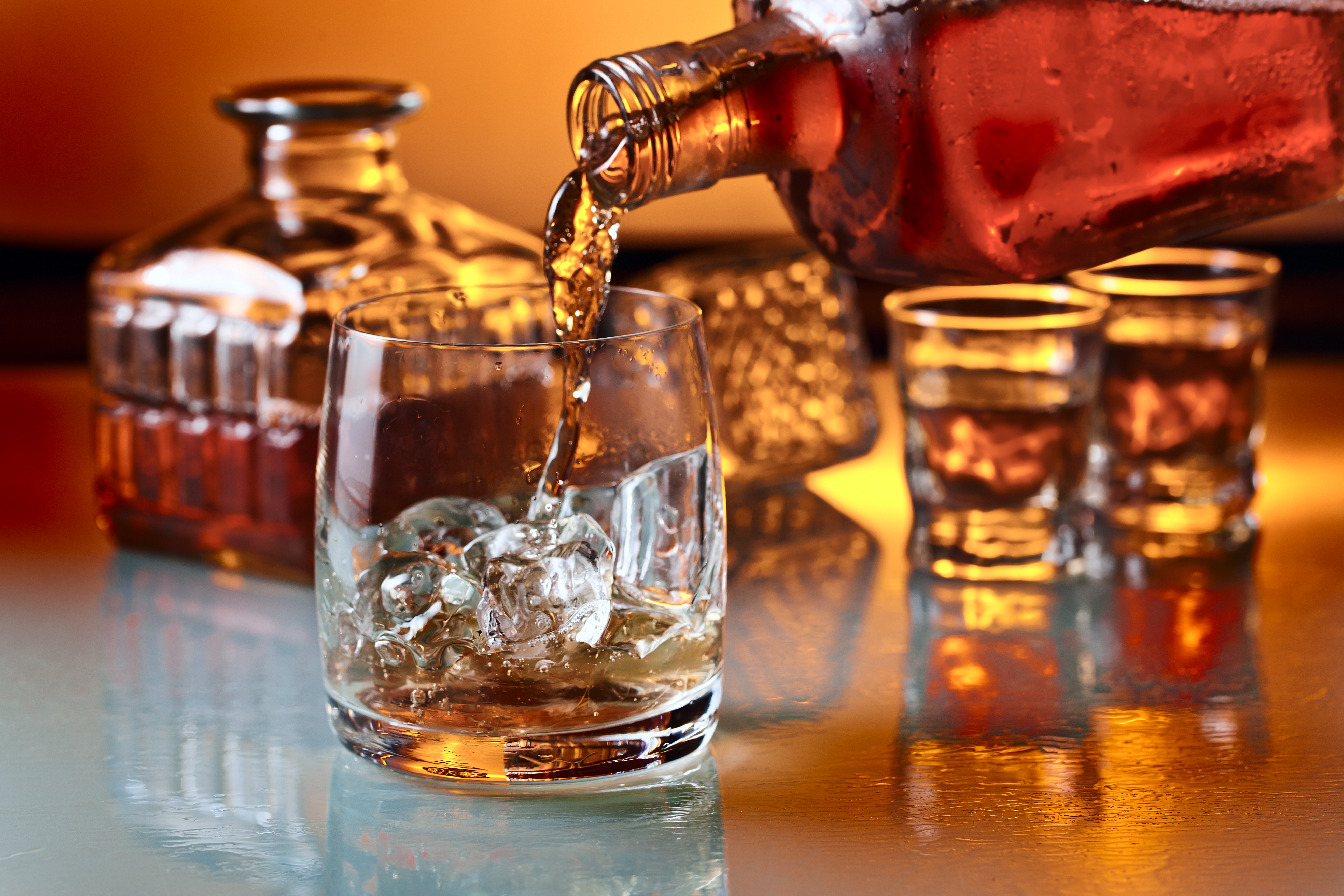 Luksuriøs gyldenbrun whiskey der hældes over isklumper i et krystalglas, med dekanterflasker og shotglas på et elegant glasbord