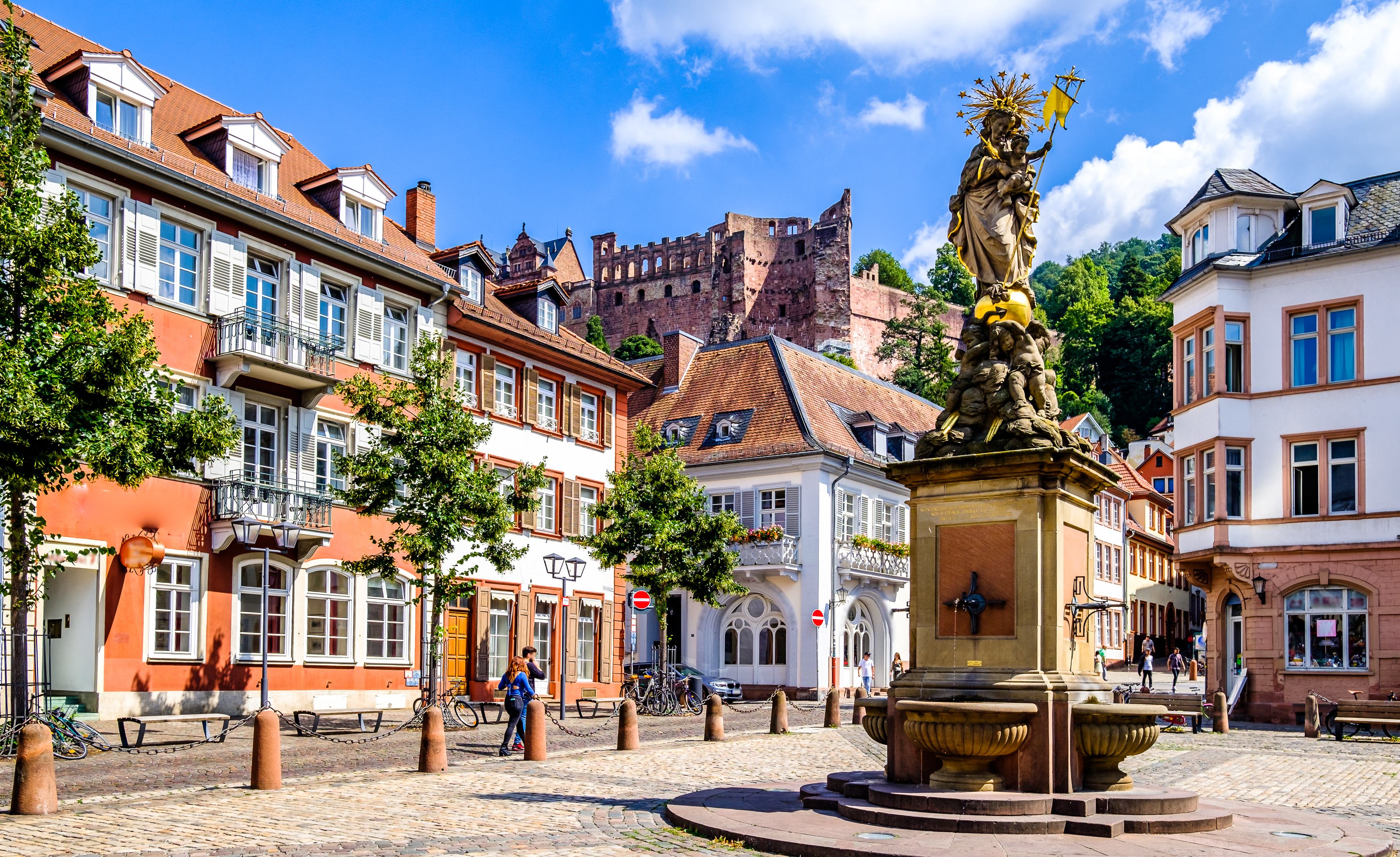 Pittoresk torv i Heidelberg med farverige bygninger, barokt springvand og udsigt til slotsruinerne på bakkerne over den historiske tyske by