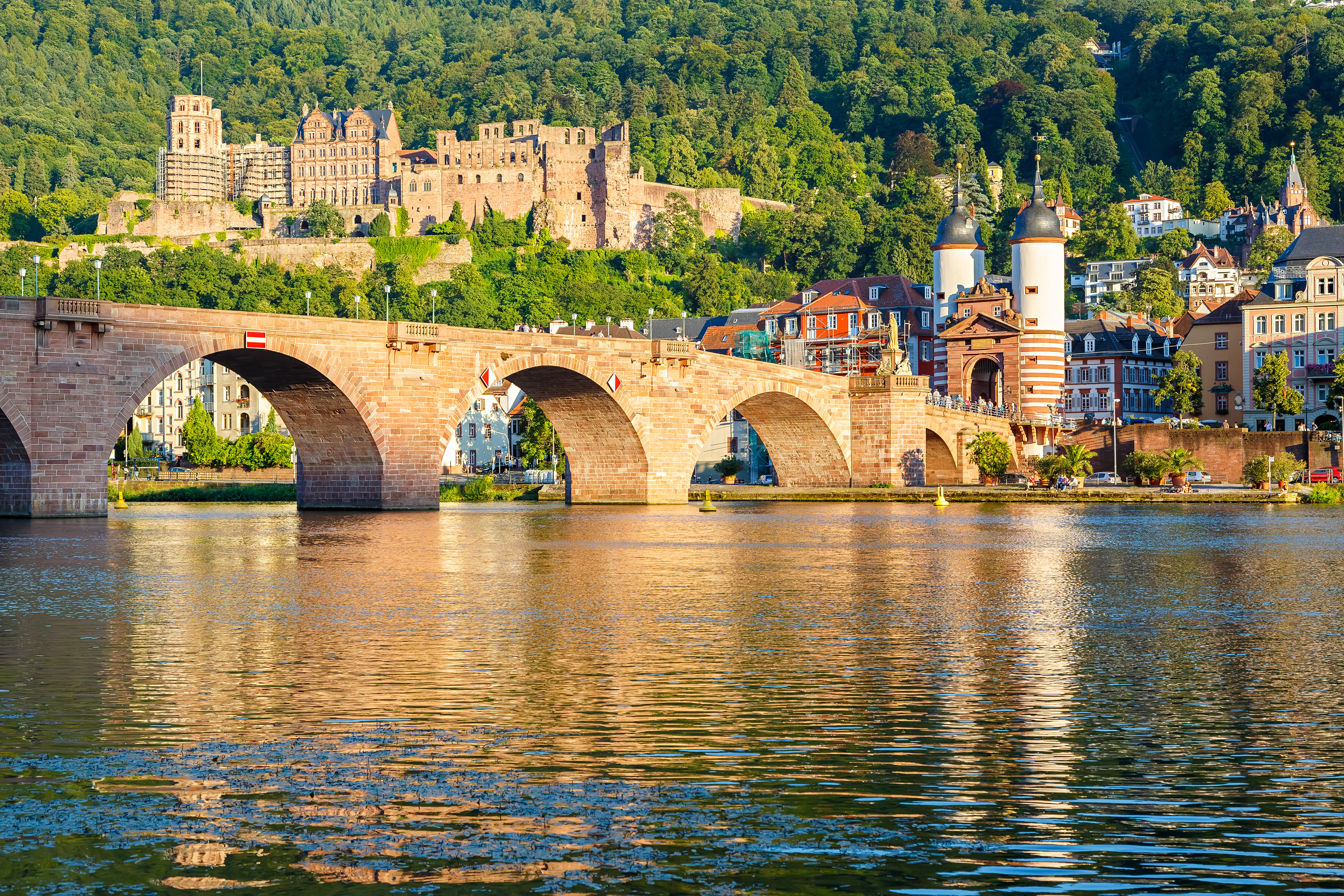 Historisk stenbro over Neckar-floden i Heidelberg med udsigt til slotsruiner og frodig skovbakke i baggrunden