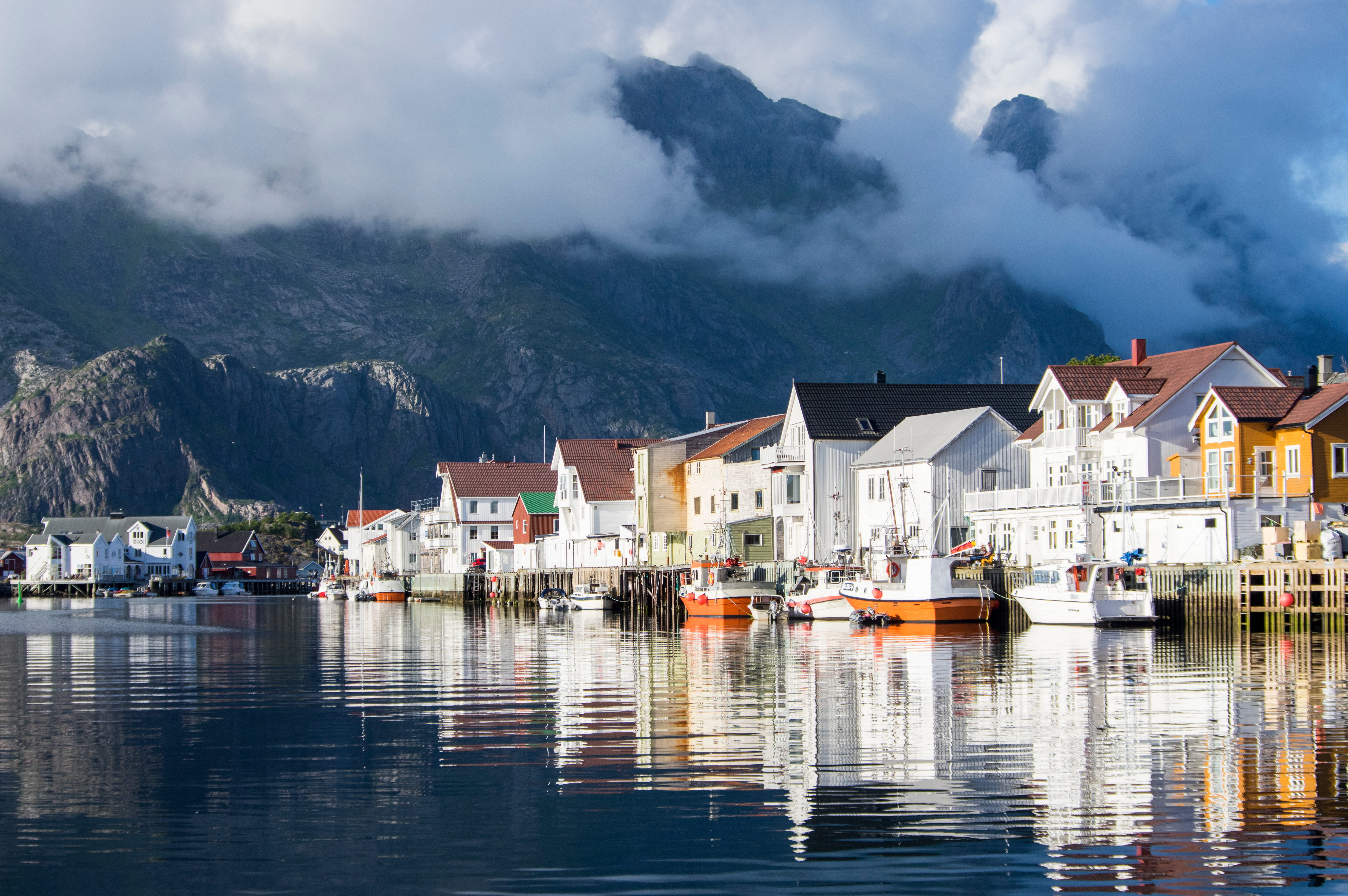 Henningsvær fiskerihavn på Lofoten om sommeren med roligt havnevand, farverige huse og dramatiske skydækkede bjerge i Norge