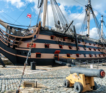 Det ikoniske britiske krigsskib HMS Victory fortøjet ved Portsmouth Historic Dockyard med Union Jack vajende fra masten og en gammel kanon i forgrunden