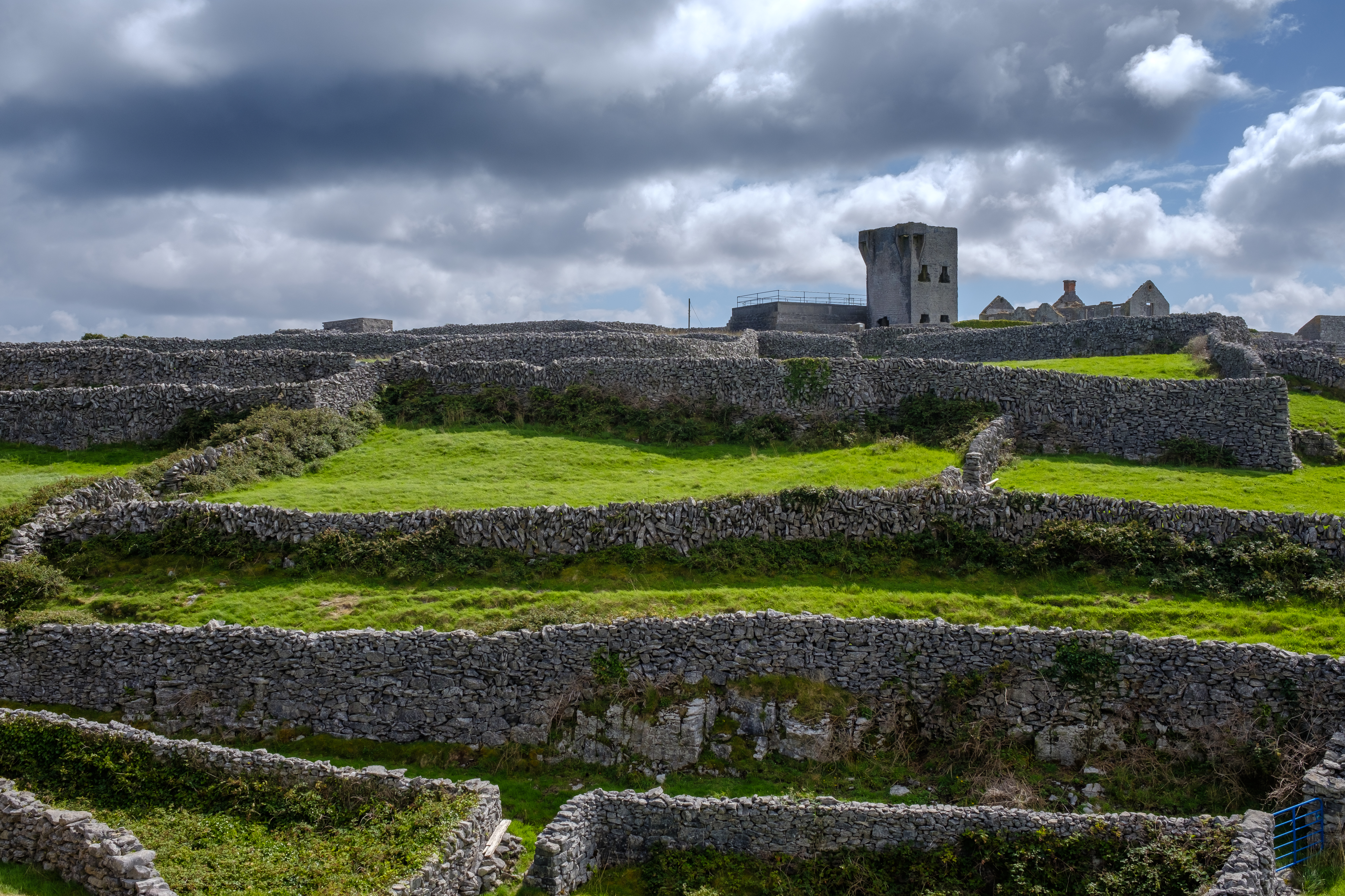 Udsigt over Inisheer på Aran-øerne med traditionelle stenvolde, frodige grønne marker og et gammelt tårn under en dramatisk skyet irsk himmel