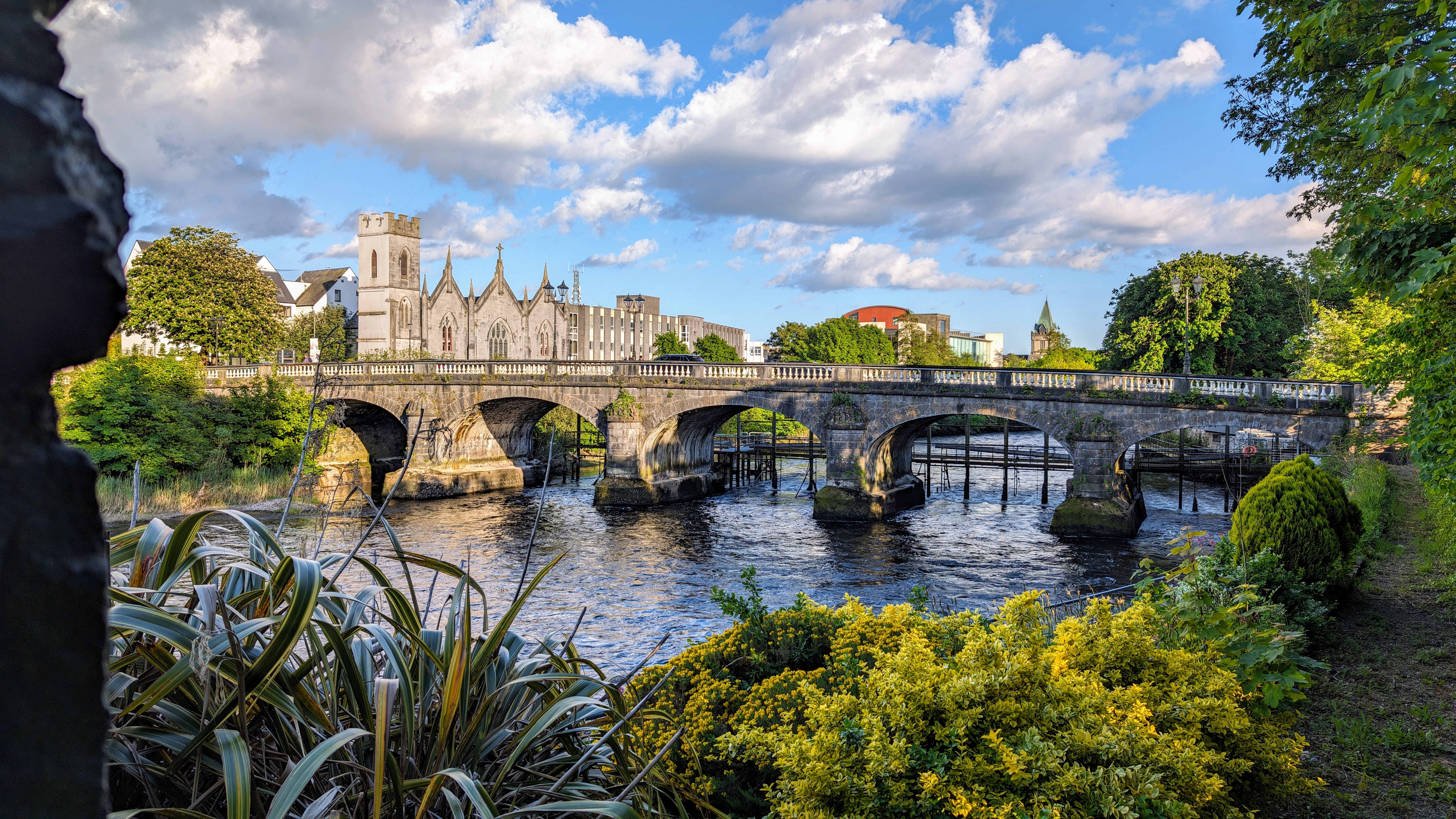 Idyllisk sceneri ved Salmon Weir broen i Galway, der forbinder katedralen med retsbygningen over floden Corrib med grønne områder i forgrunden