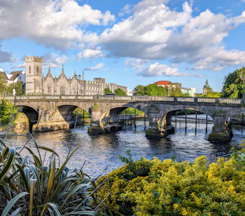 Idyllisk sceneri ved Salmon Weir broen i Galway, der forbinder katedralen med retsbygningen over floden Corrib med grønne områder i forgrunden