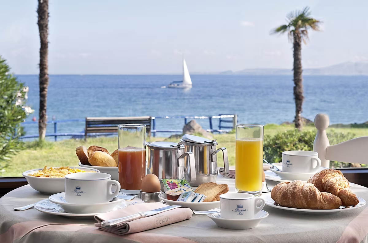 Feriemorgenmad med kaffe, juice og croissanter med udsigt til Middelhavet og en sejlbåd fra Ischia
