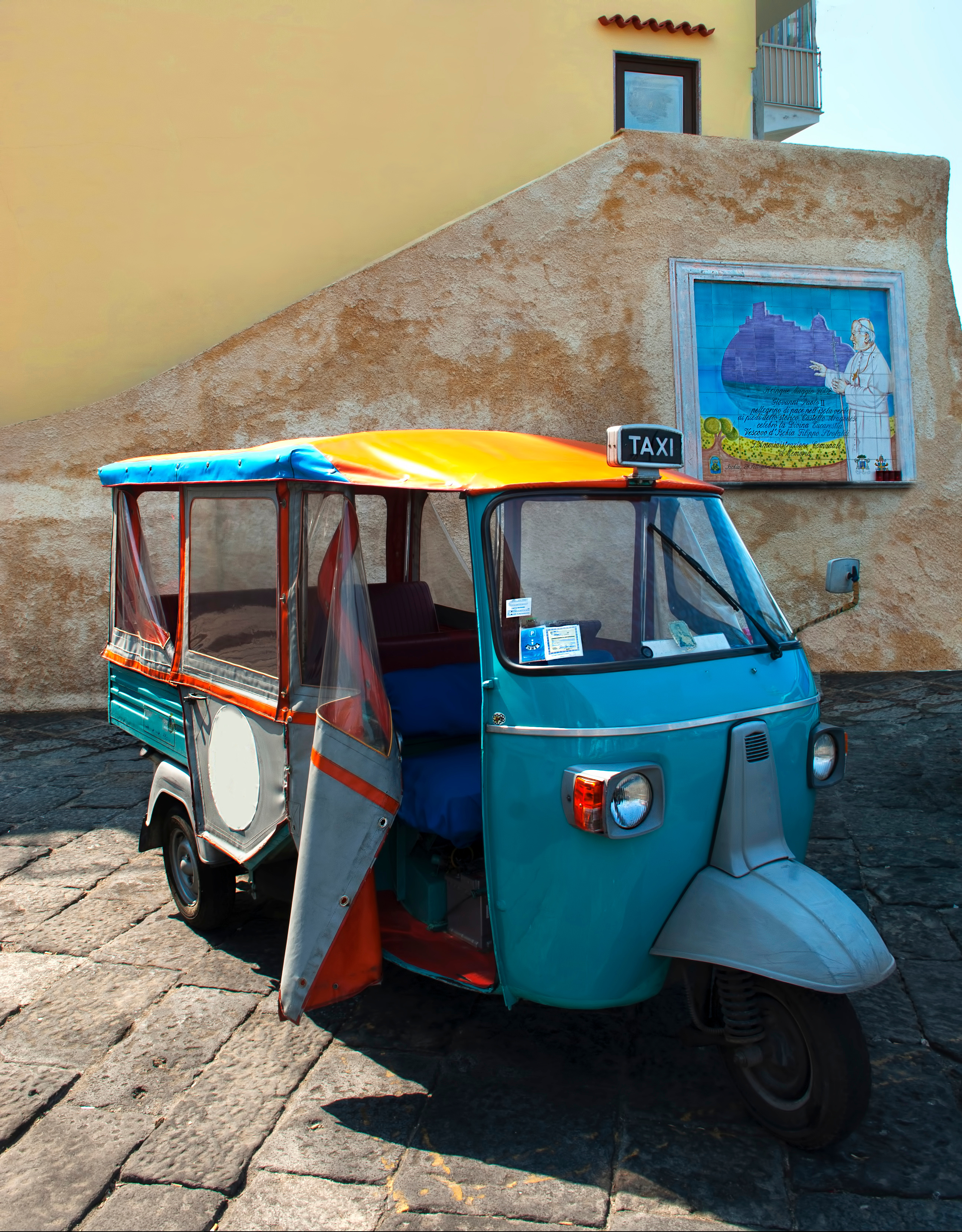 Charmerende farverig taxa på brolagt plads i Ischia Porto med autentisk italiensk gadebillede og dekorativ vægmosaik