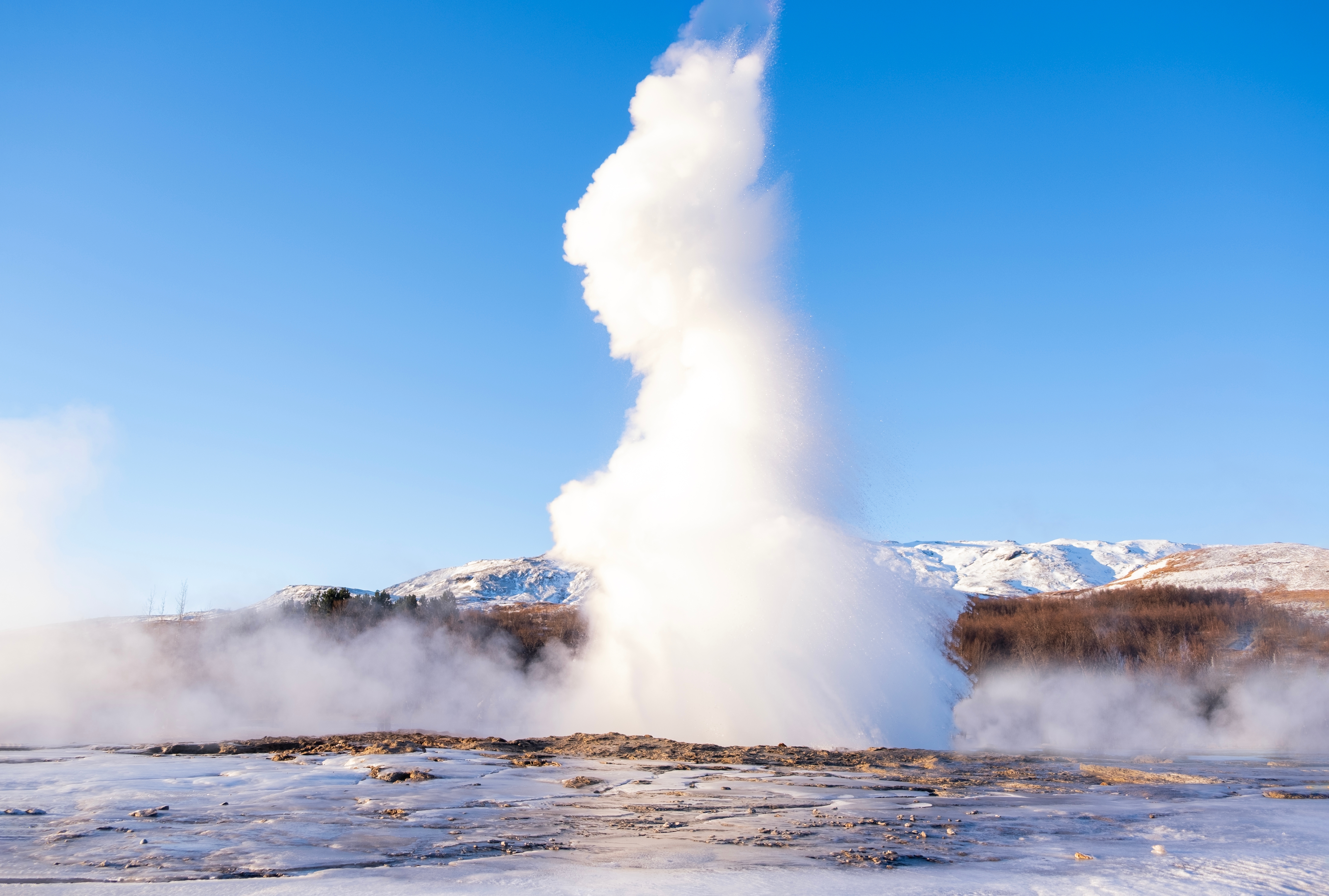 Kraftigt udbrud fra Strokkur geyseren på Island med snedækket terræn og blå himmel i baggrunden