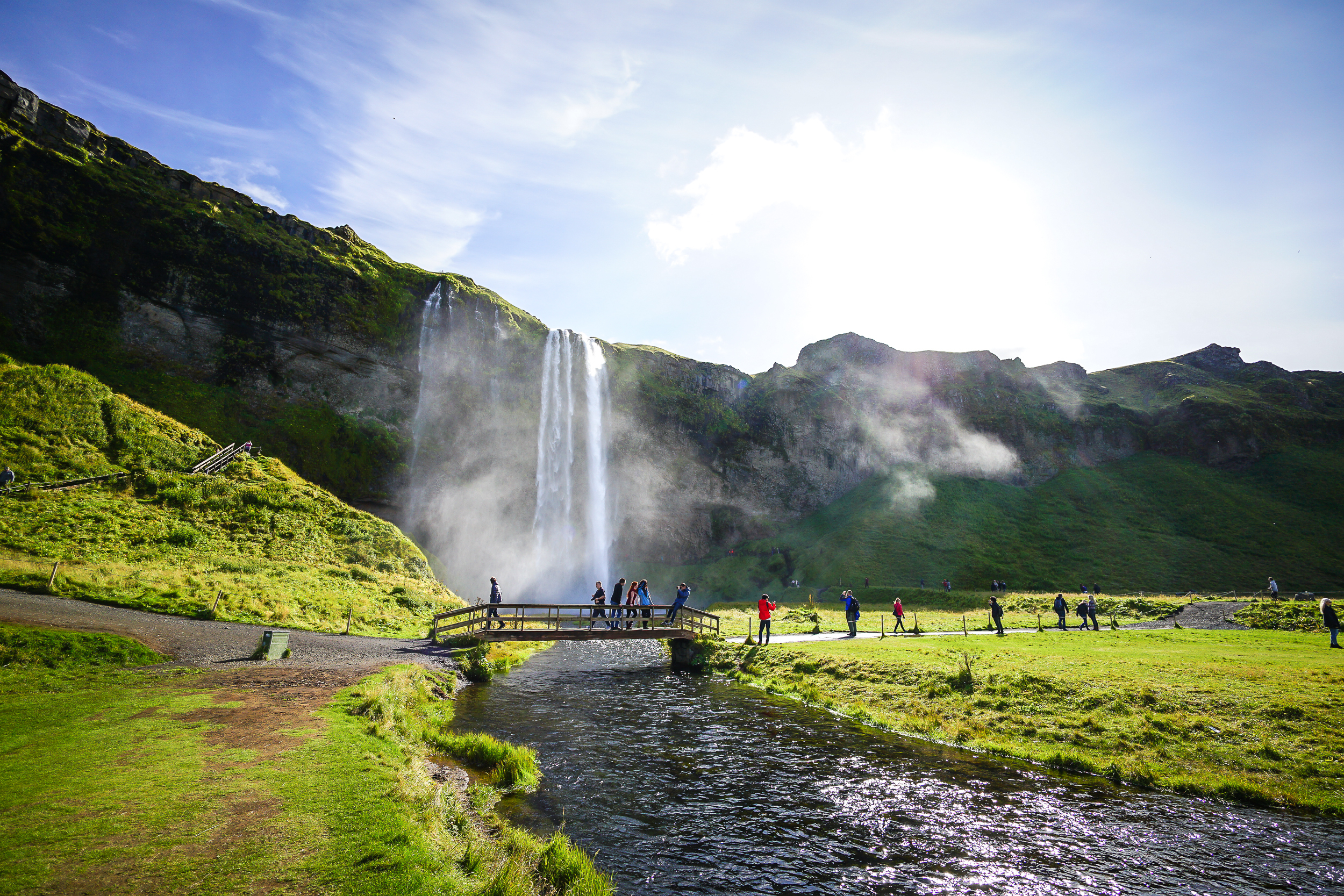 Turister nyder udsigten til Islands berømte Seljalandsfoss vandfald fra en træbro over den klare flod omgivet af grønne marker
