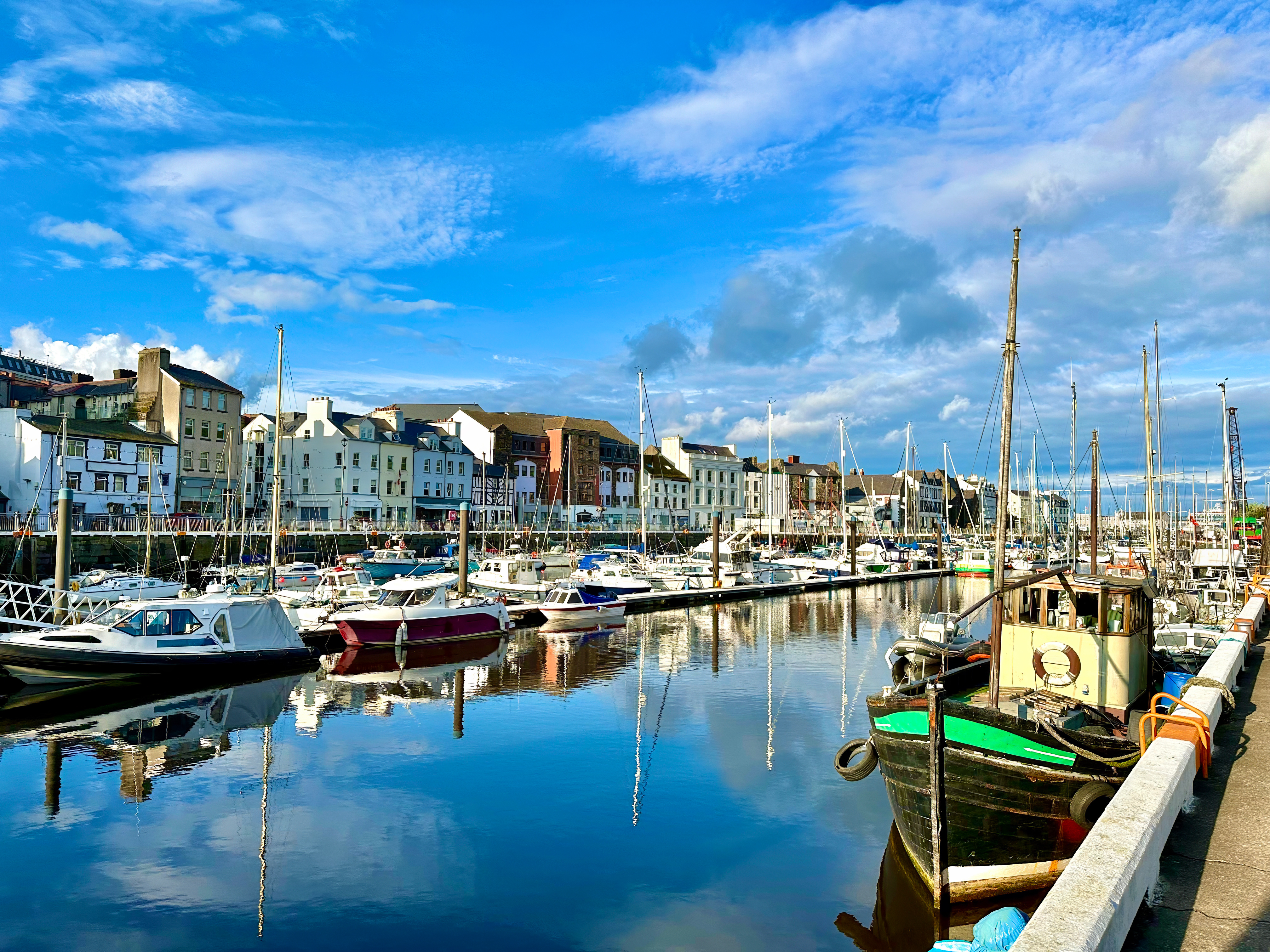 Solrig dag ved Douglas Marina på Isle of Man med sejlbåde og lystfartøjer fortøjet langs kajen og charmerende bygninger i baggrunden