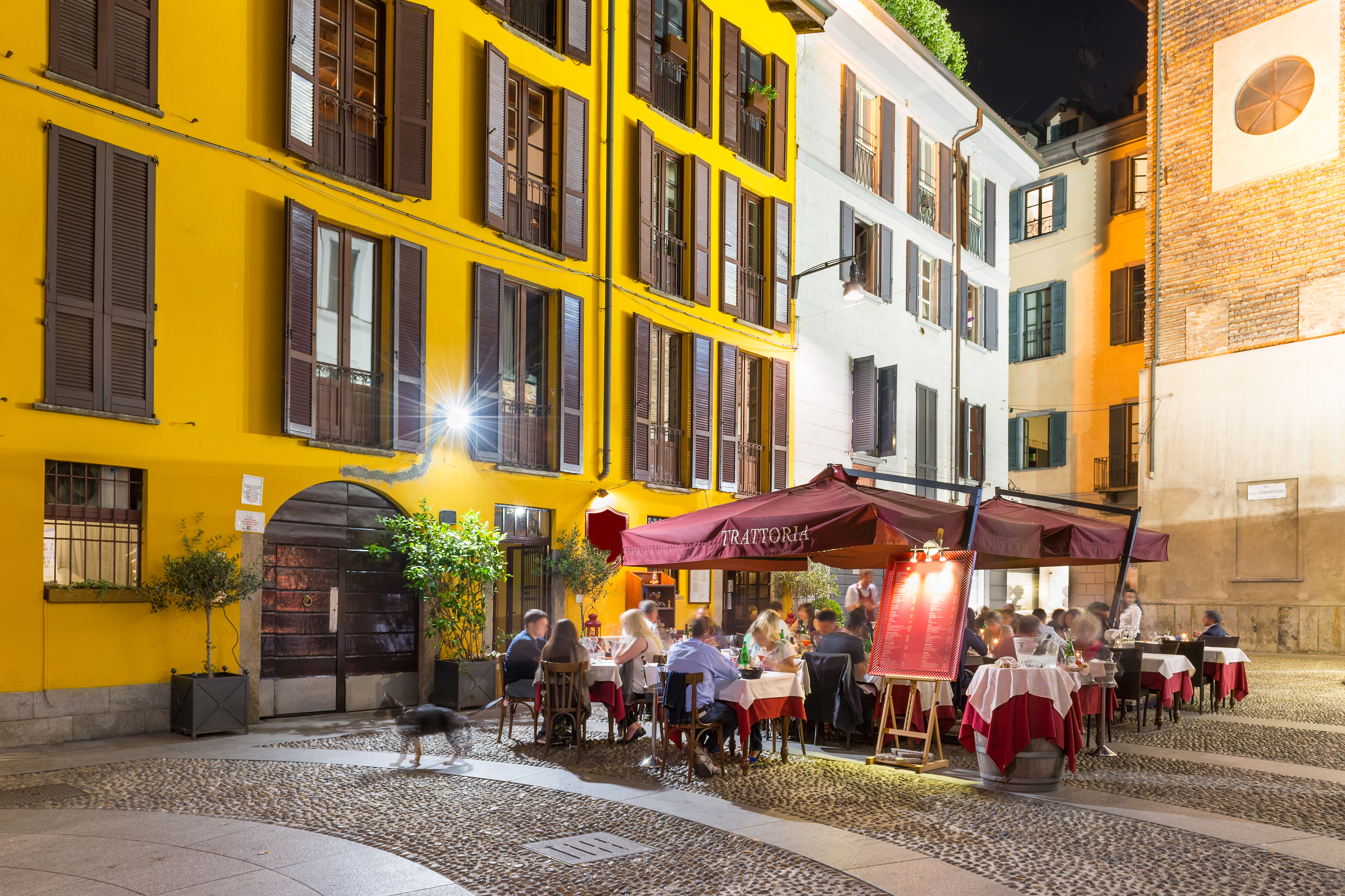 Idyllisk italiensk trattoria med udendørs servering på en brostensbelagt gade i Milano om aftenen med gæster ved borde med rød-hvide duge