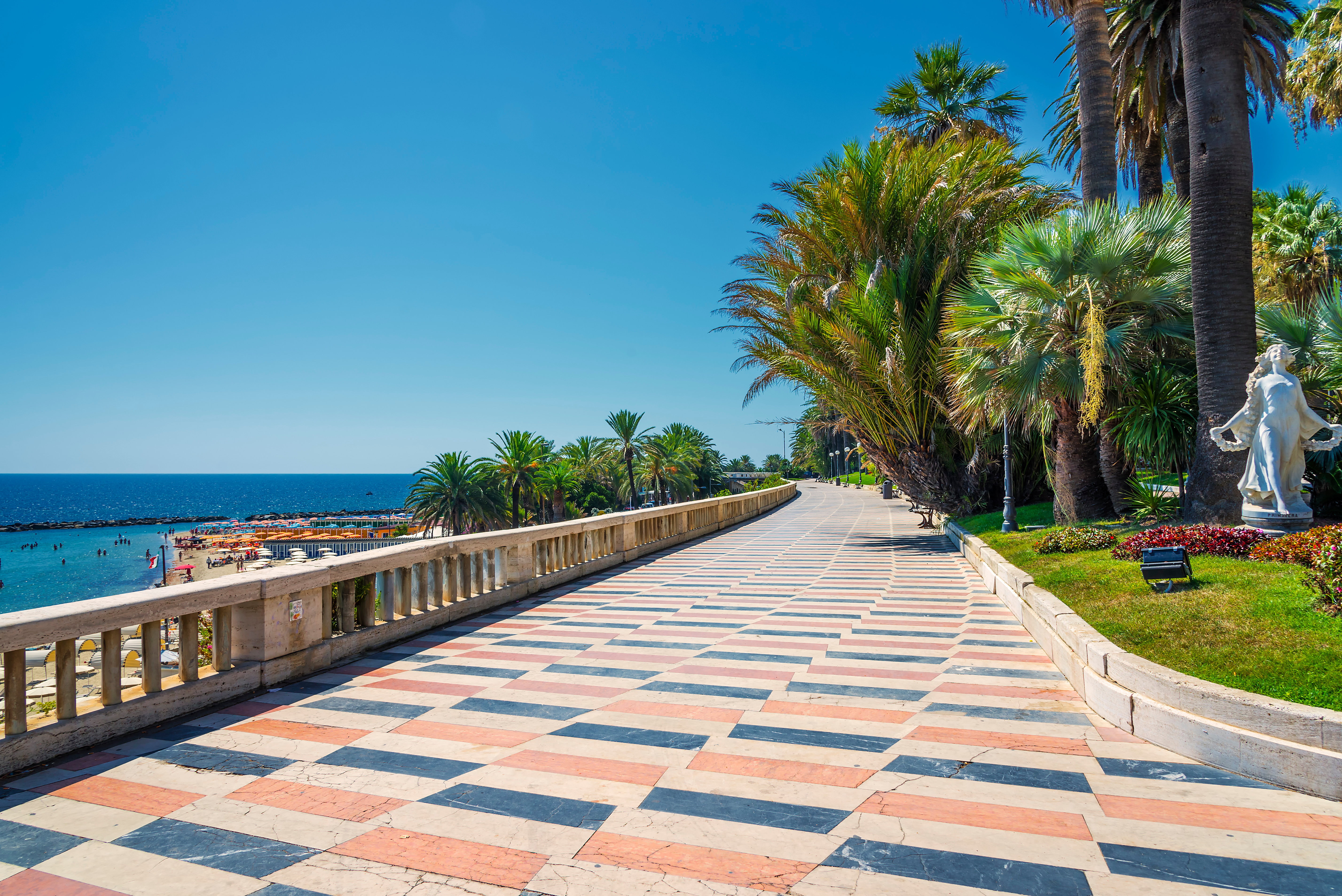 Promenaden i San Remo på den italienske riviera med hvid klassisk statue, palmer og stranden i baggrunden på en solrig dag