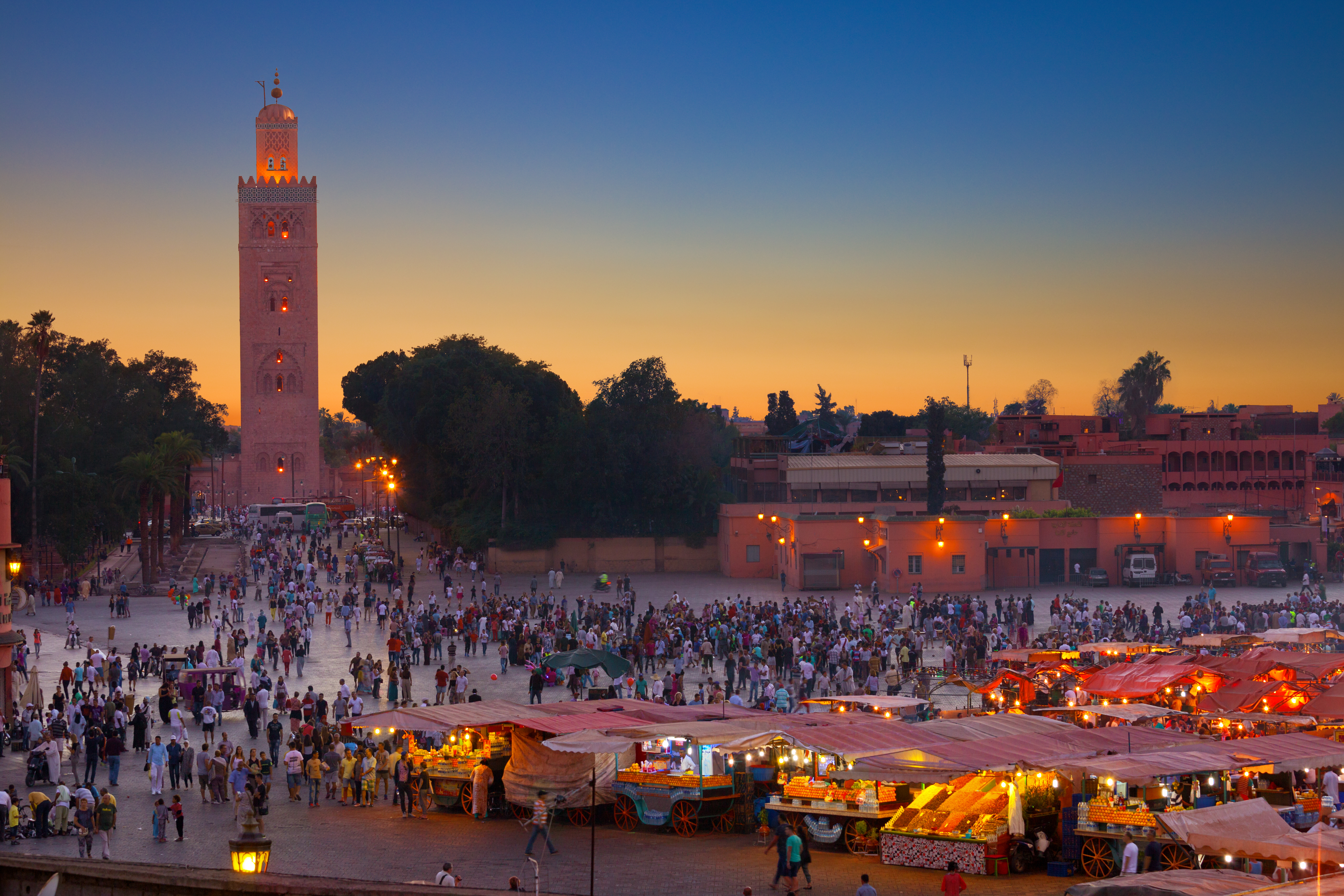 Livlig Jemaa el-Fna plads i Marrakech med tusindvis af mennesker, markedsboder og den ikoniske Koutoubia-minaret ved solnedgang