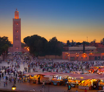 Livlig Jemaa el-Fna plads i Marrakech med tusindvis af mennesker, markedsboder og den ikoniske Koutoubia-minaret ved solnedgang