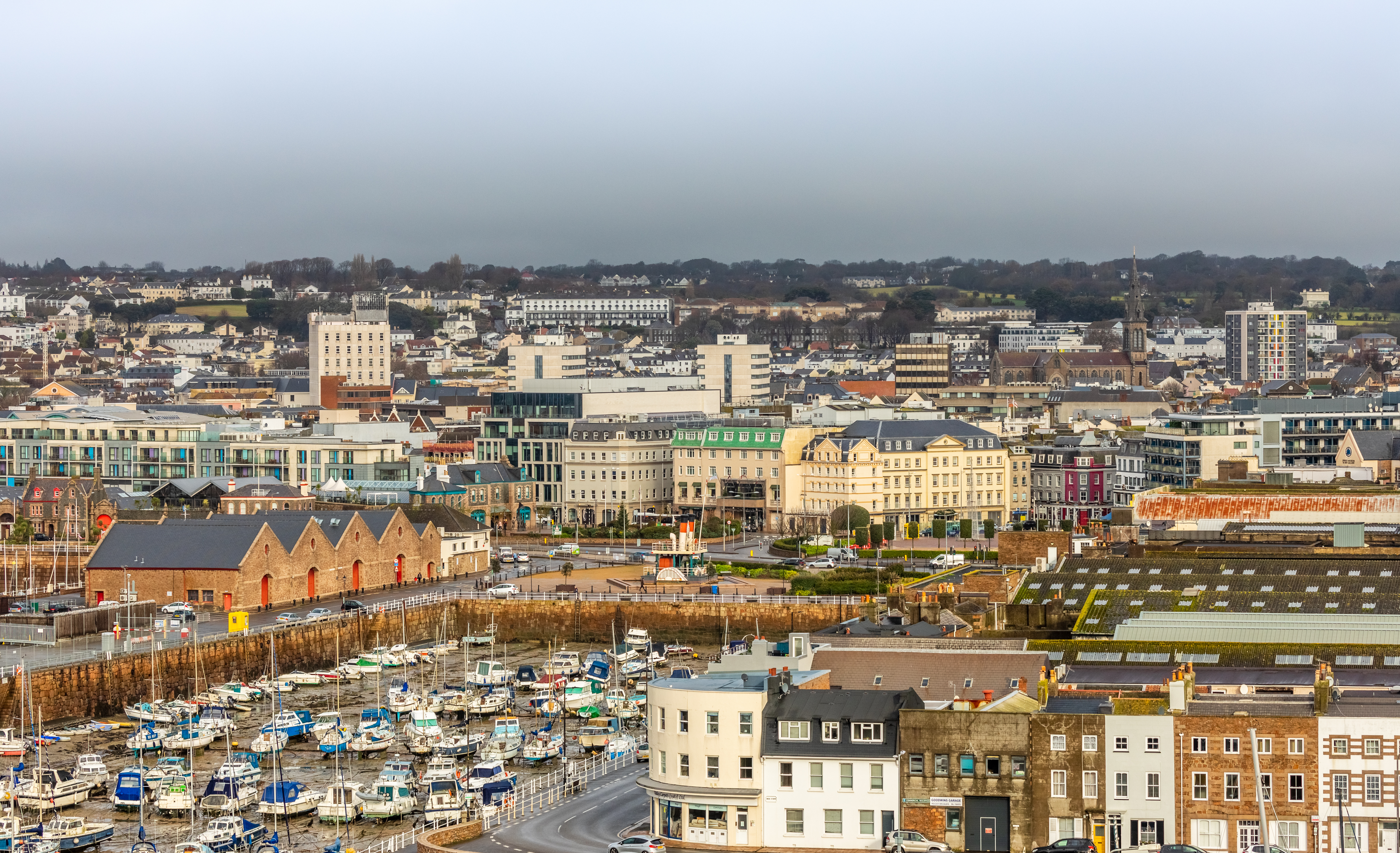 Panoramabillede af Saint Helier på Jersey med den livlige marina, historiske bygninger og grønne bakkelandskab i horisonten på en overskyet dag
