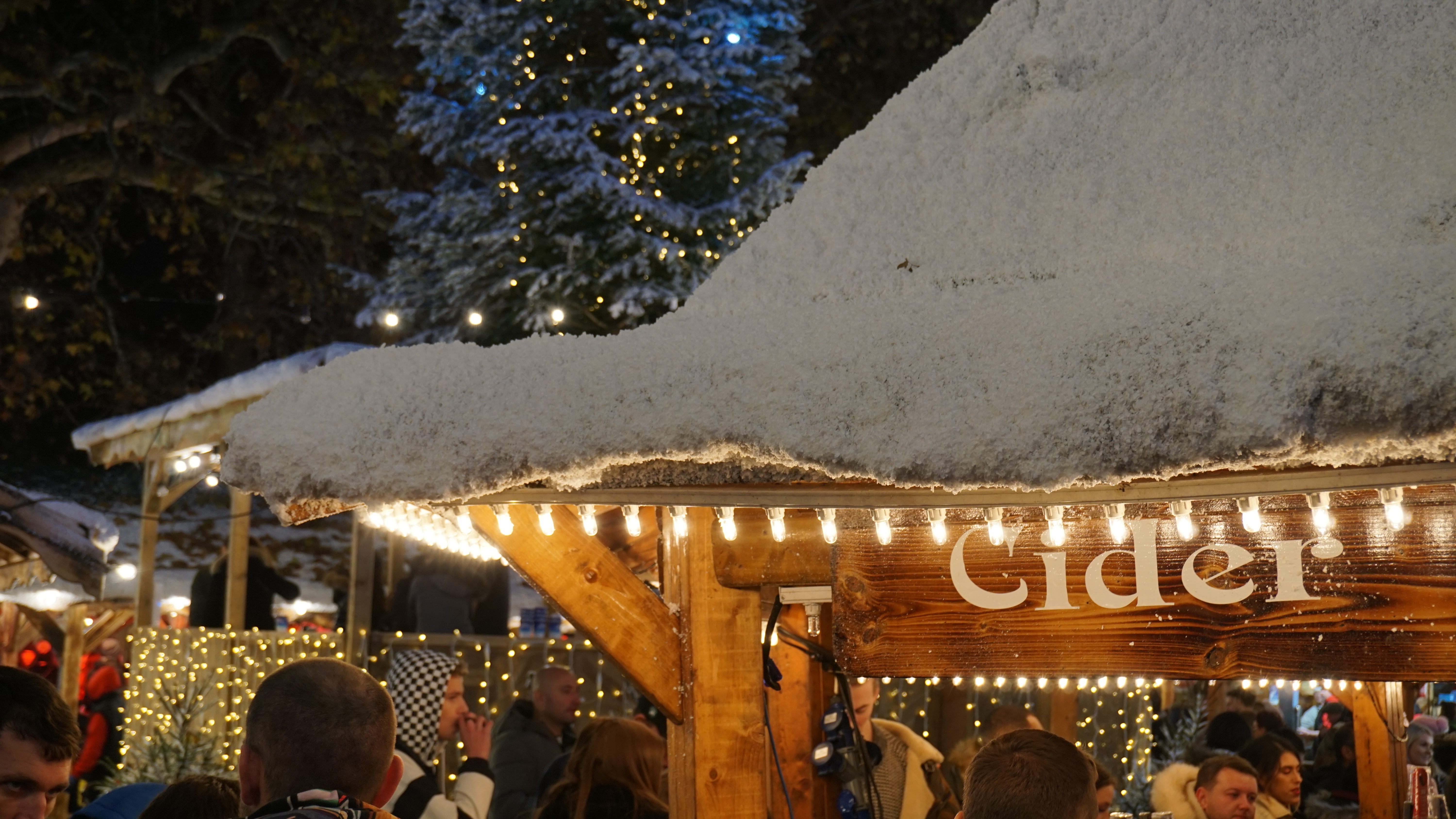 Julemarked i London om natten med sne på tagene, en oplyst cider-bod og et smukt juletræ med lys i baggrunden