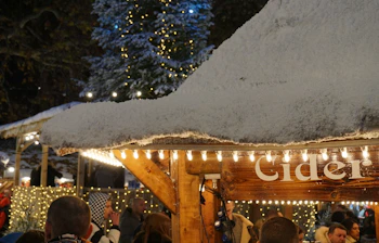 Julemarked i London om natten med sne på tagene, en oplyst cider-bod og et smukt juletræ med lys i baggrunden