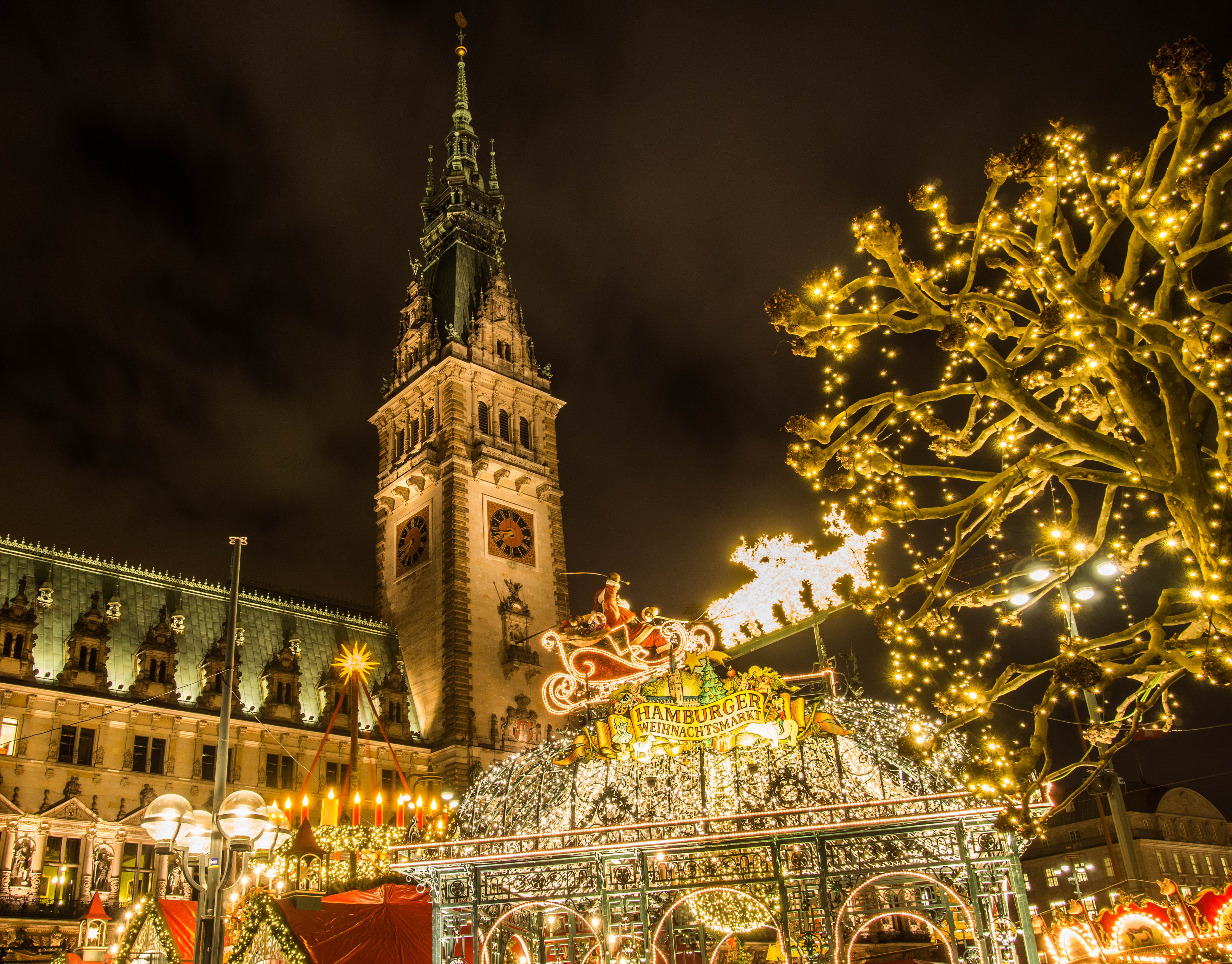 Idyllisk julestemning på Hamburgs traditionsrige julemarked, hvor de festlige boder og dekorationer skaber en magisk atmosfære foran det historiske rådhus i aftenlys