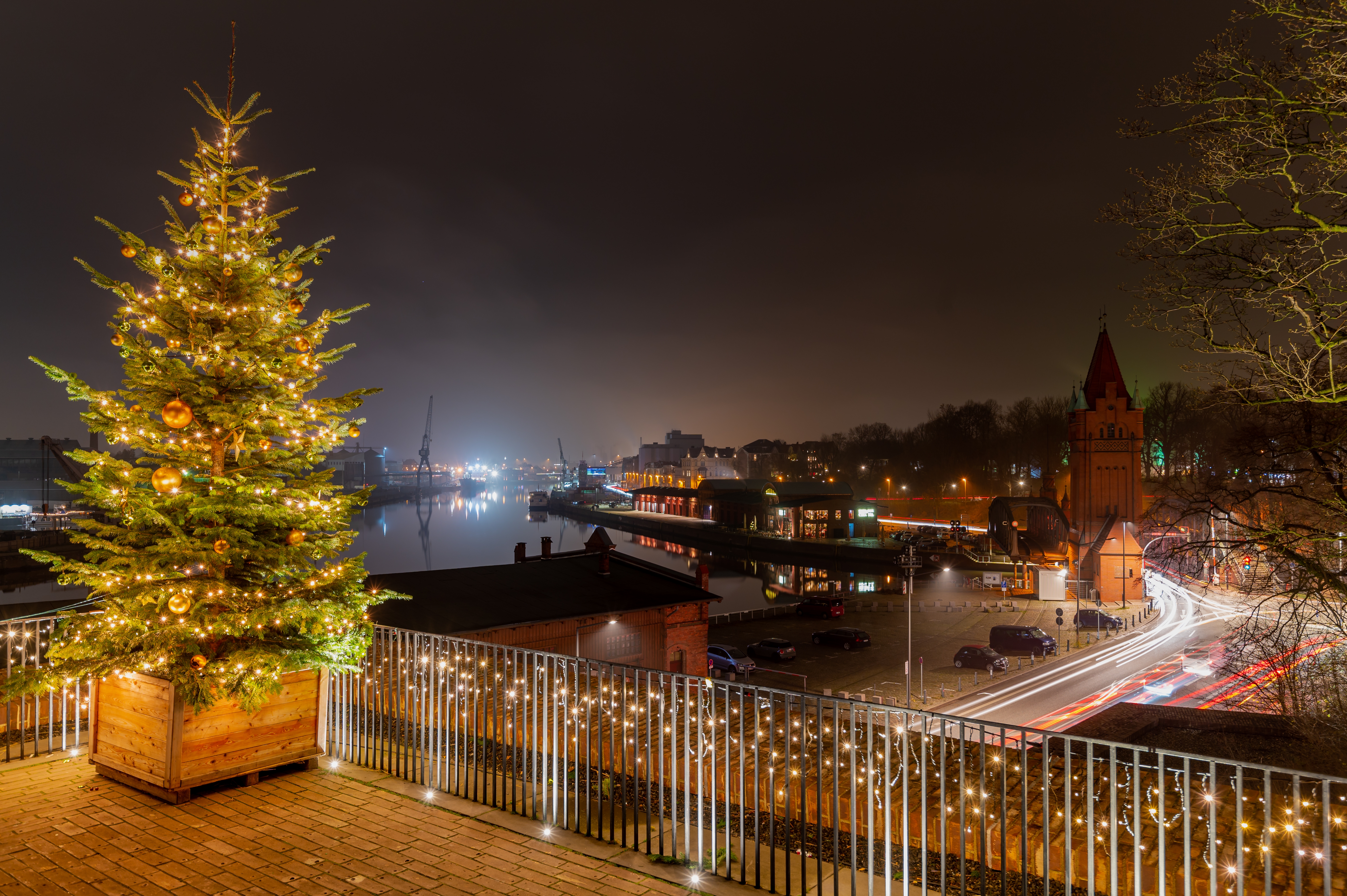 Festligt oplyst juletræ ved den hanseatiske by Lübecks havn om natten, med middelalderlige murstenstårne og den rolige flod Trave der skaber en magisk juleatmosfære
