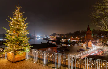 Festligt oplyst juletræ ved den hanseatiske by Lübecks havn om natten, med middelalderlige murstenstårne og den rolige flod Trave der skaber en magisk juleatmosfære