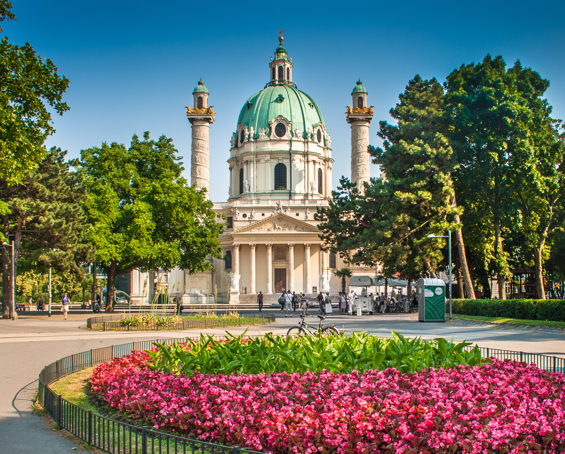 Wiens ikoniske Karlskirche med grøn kuppel og barokfacade omgivet af frodig park med farverige blomster i sommersolen