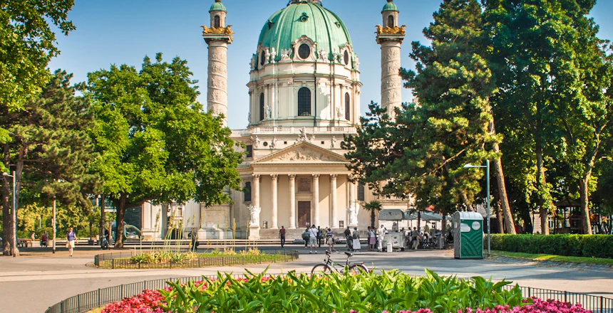 Wiens ikoniske Karlskirche med grøn kuppel og barokfacade omgivet af frodig park med farverige blomster i sommersolen