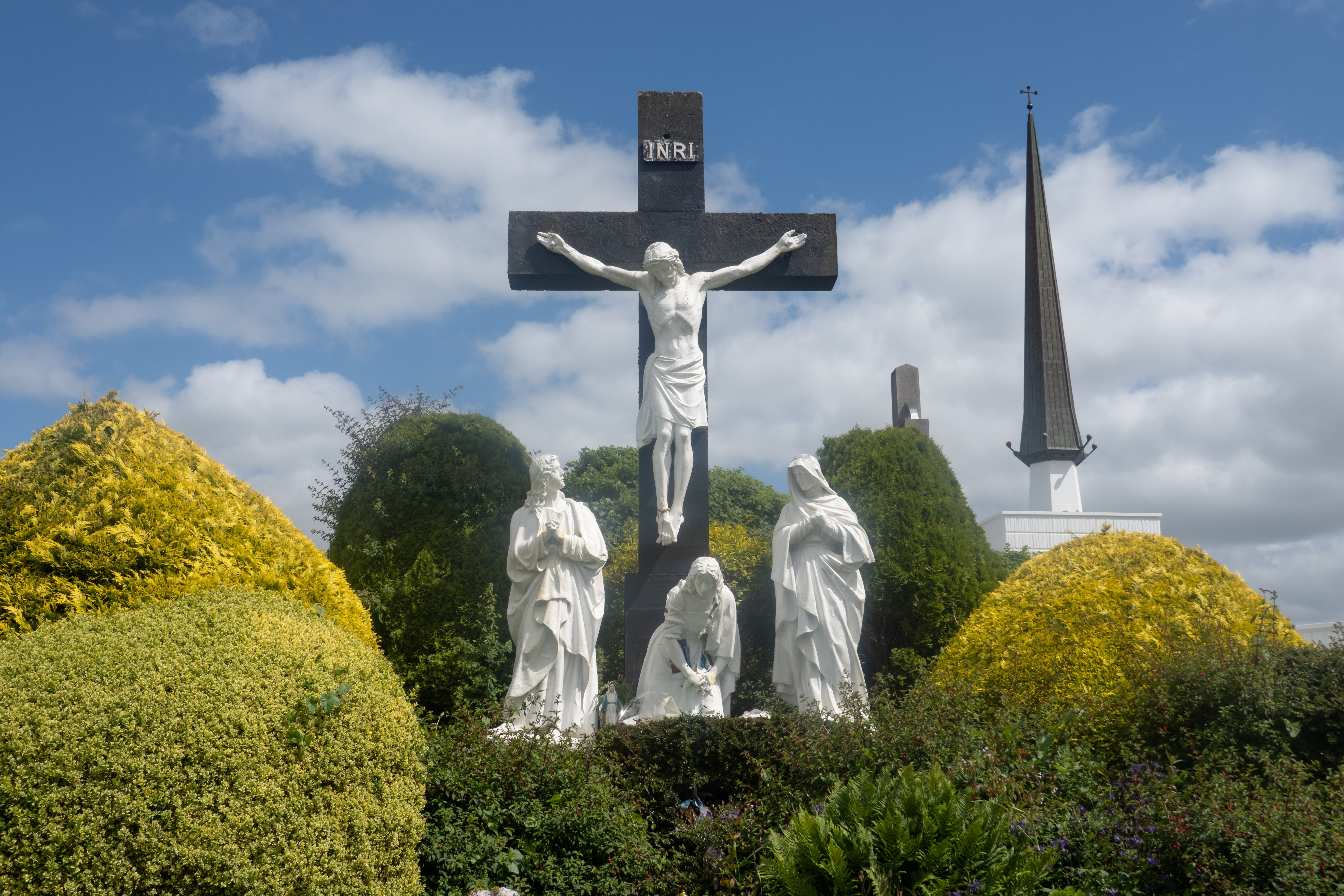 Smukke hvide marmorstatuer ved Knock Shrine i Irland forestiller korsfæstelsen med Jomfru Maria og helgener, omgivet af gyldne topiarbuskadser og en kirkespir