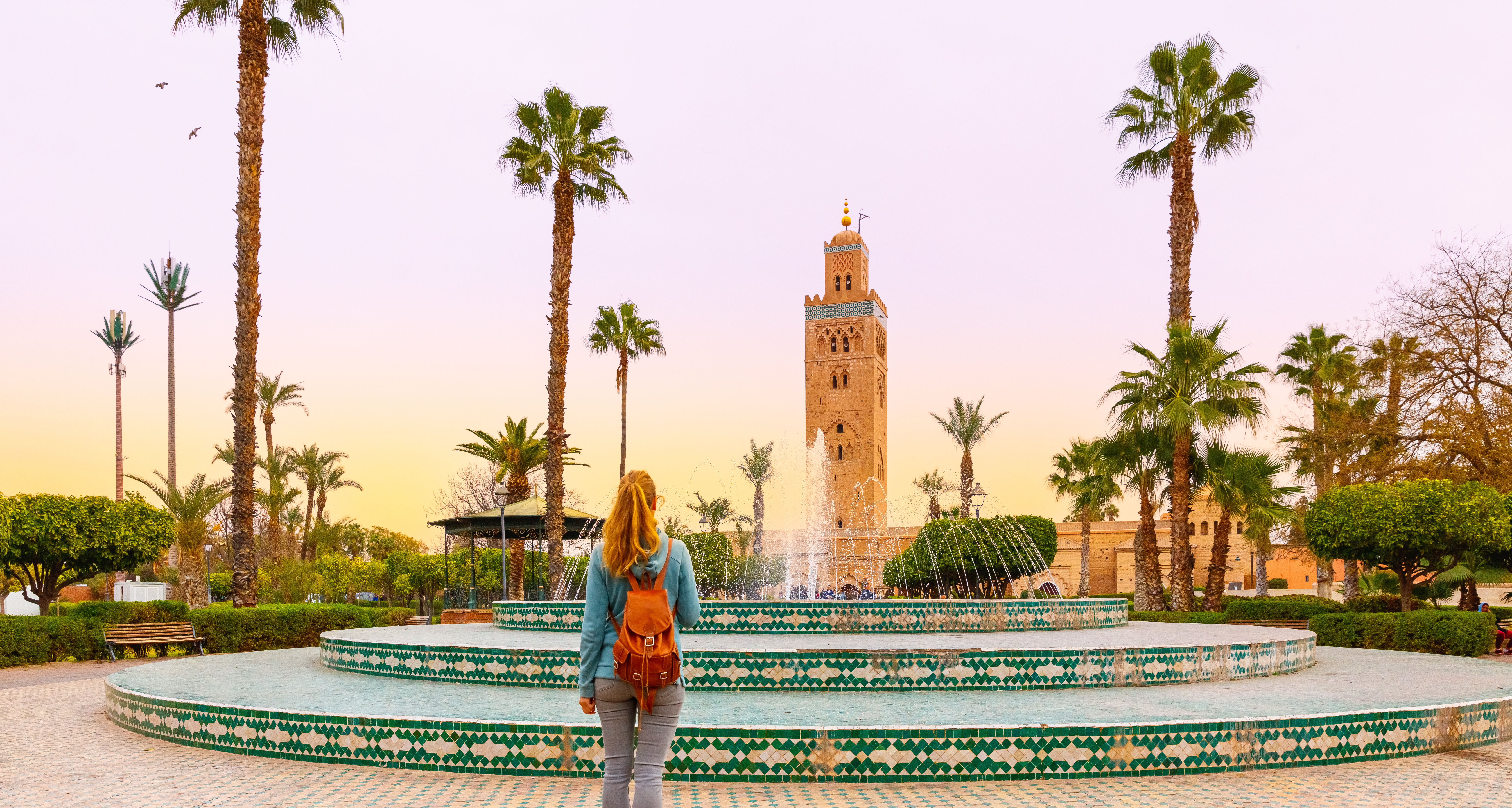 Kvindelig turist med rygsæk ser på Koutoubia-moskéens minaret ved en flot dekoreret fontæne med palmetræer i Marrakech, Marokko