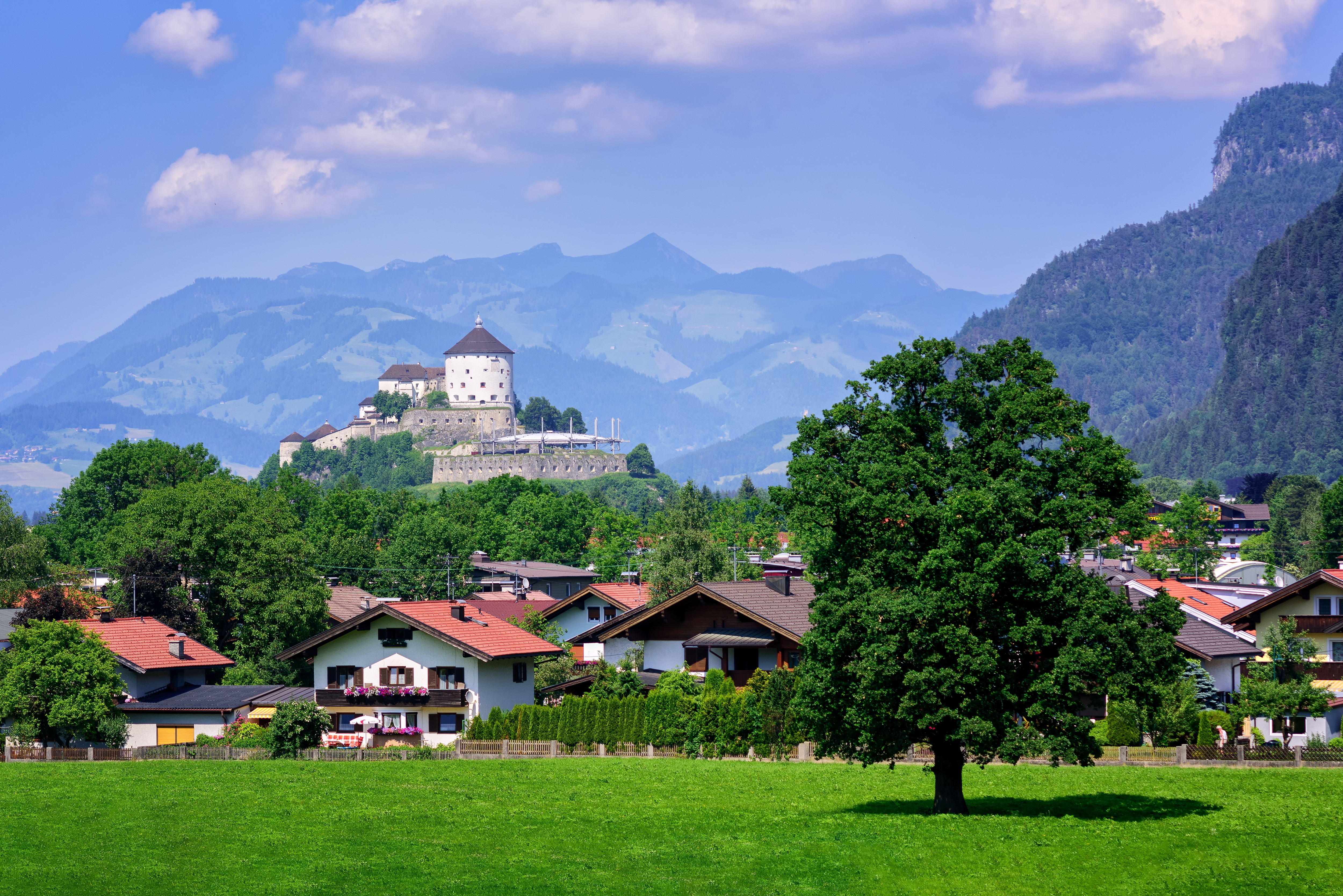 Betagende panorama over det historiske Kufstein slot på bjergtinden med den charmerende østrigske by nedenfor, omgivet af grønne enge, traditionelle alpine huse med røde tage og de majestætiske bjerge i Tyrol under blå himmel