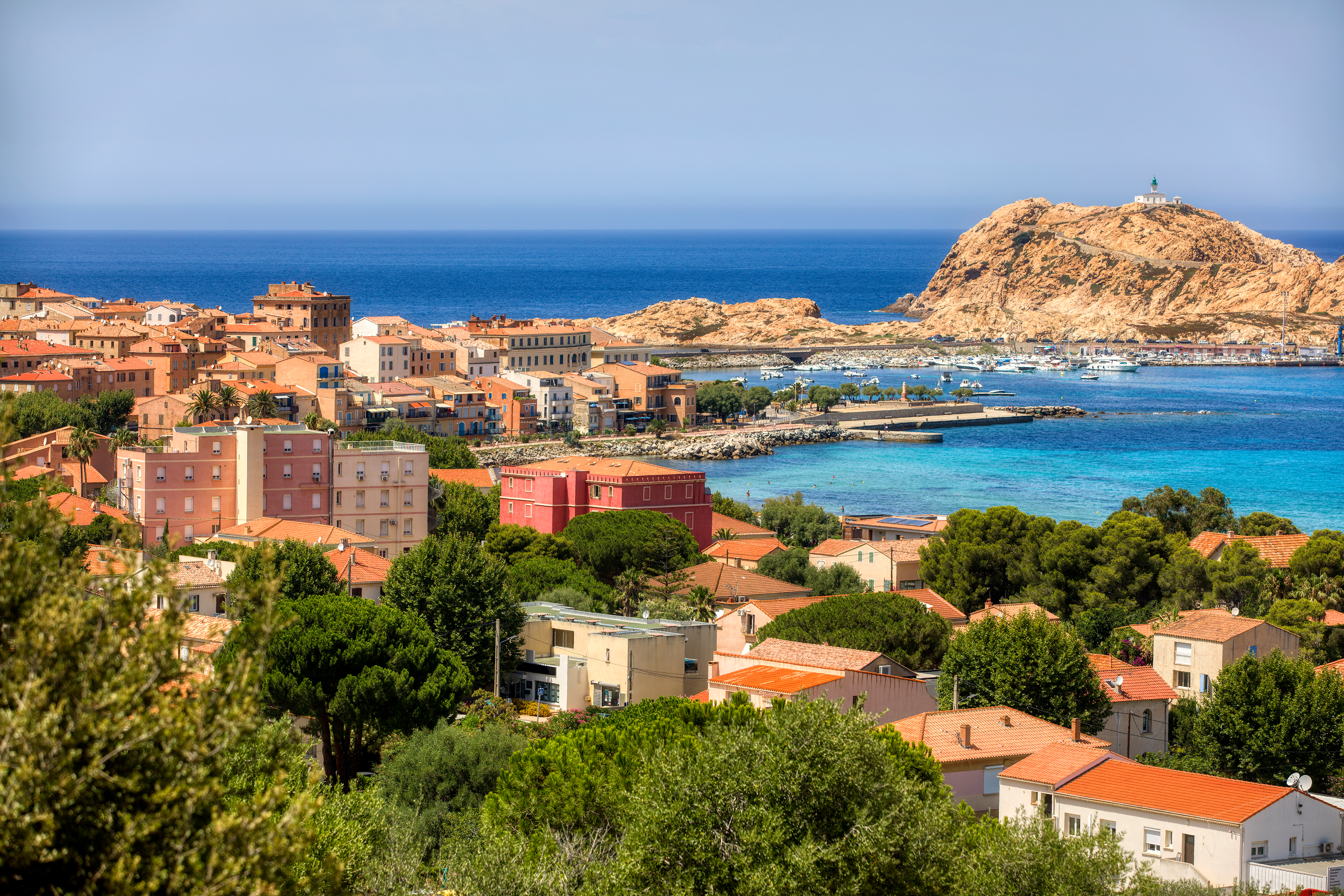 Sommerudsigt over L'Ile Rousse på Korsika med farverige huse i okker og rød, omgivet af frodige træer og det smukke turkisblå Middelhav