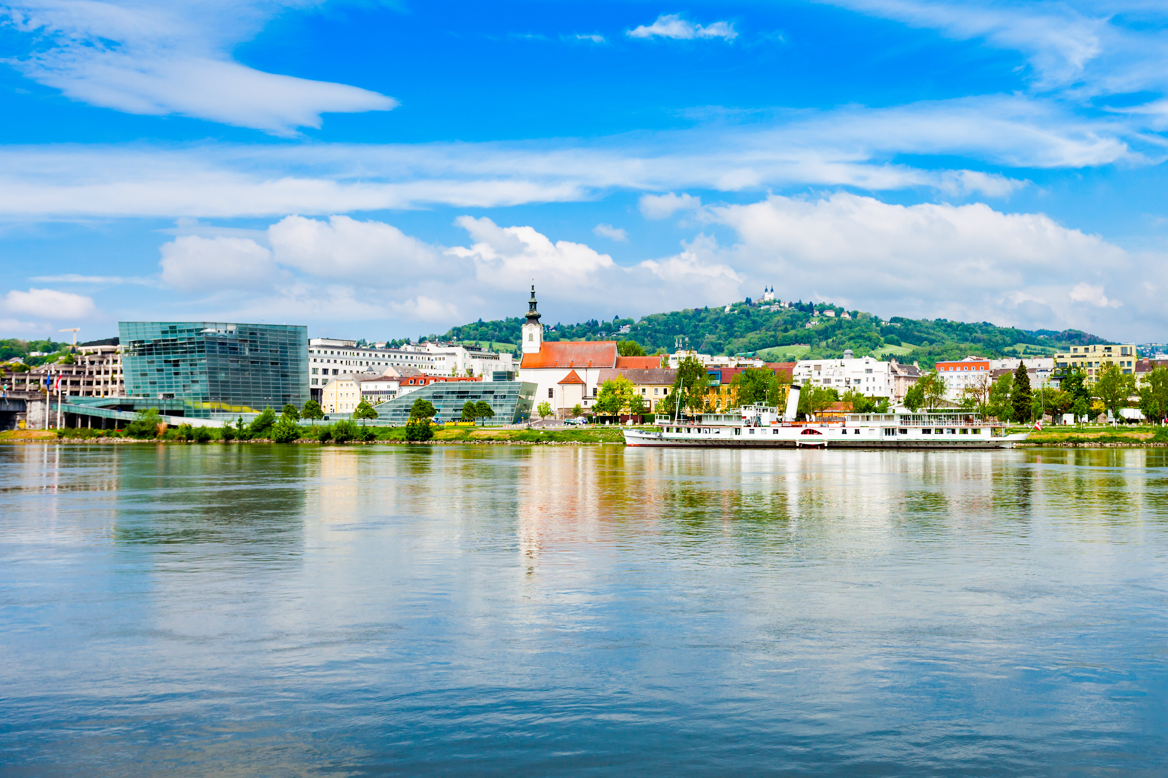 Udsigt over Linz centrum fra Donau-floden med hvidt krydstogtskib, historisk kirke og moderne glasbyggeri i Østrig