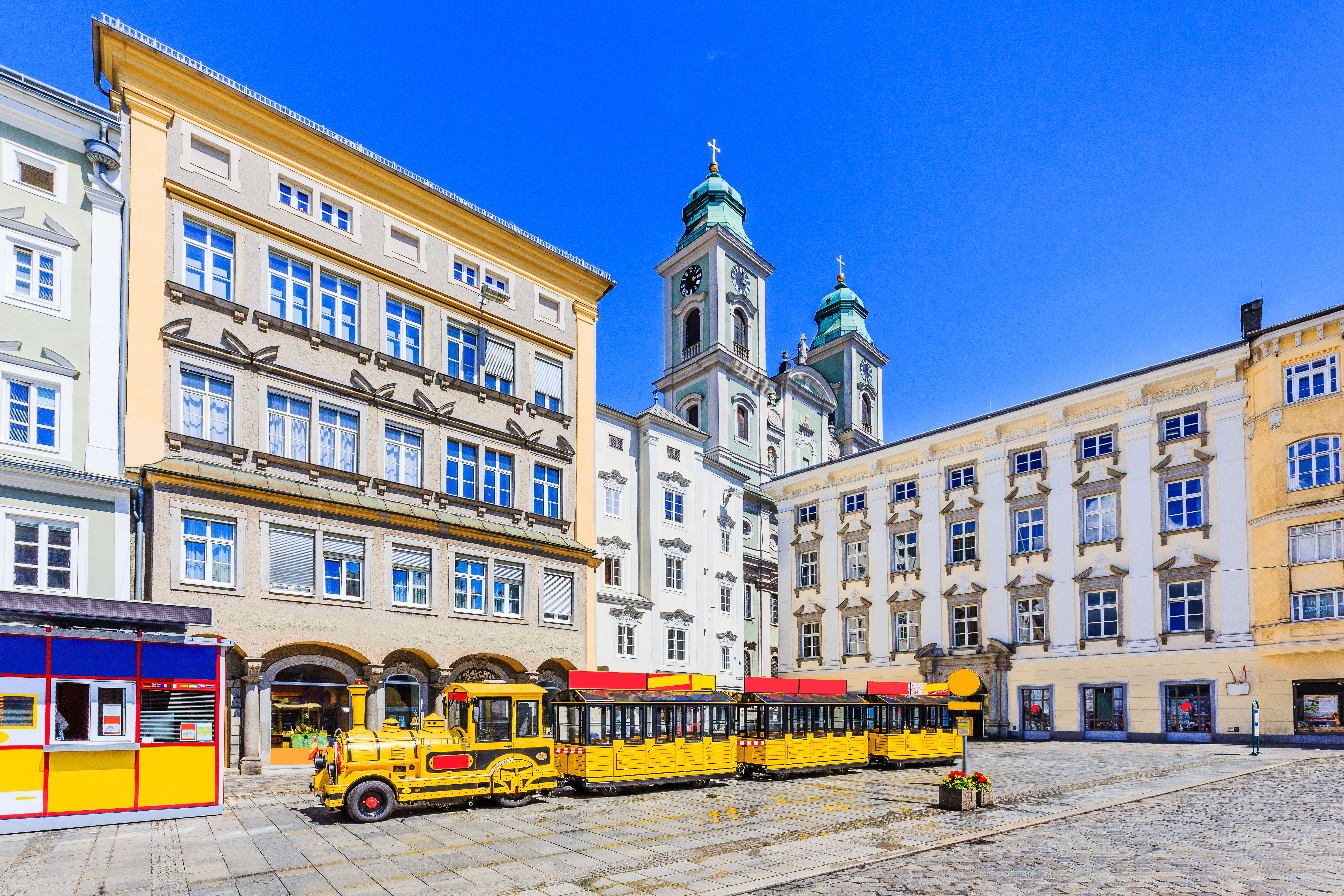 Farverig gul turisttog på Linz' smukke hovedplads med barokke bygninger og katedraltårne under blå sommerhimmel