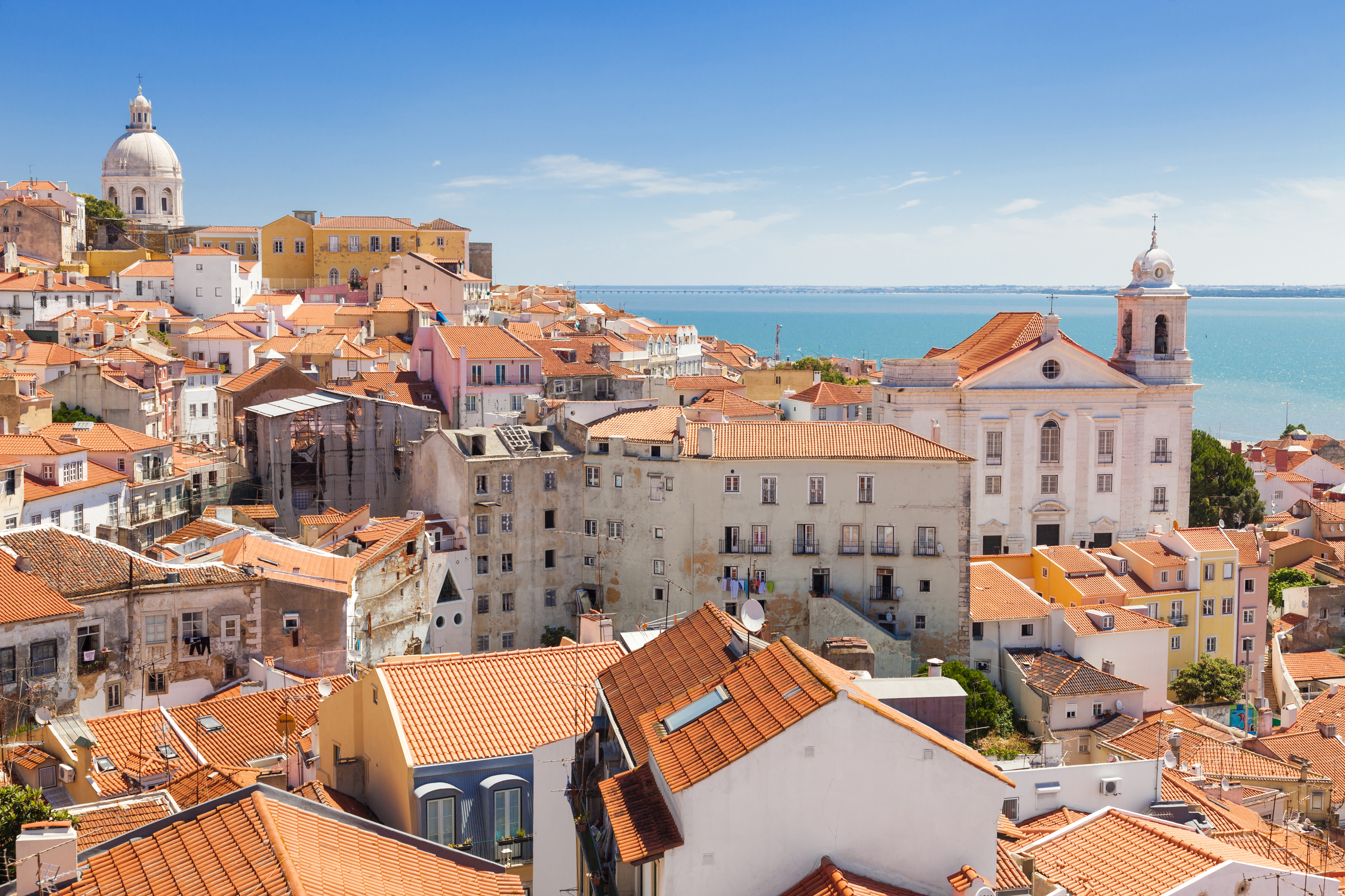 Panoramabillede af Alfama i Lissabon med tæt bebyggelse, røde tagsten og den smukke Tejo-flod bag byen