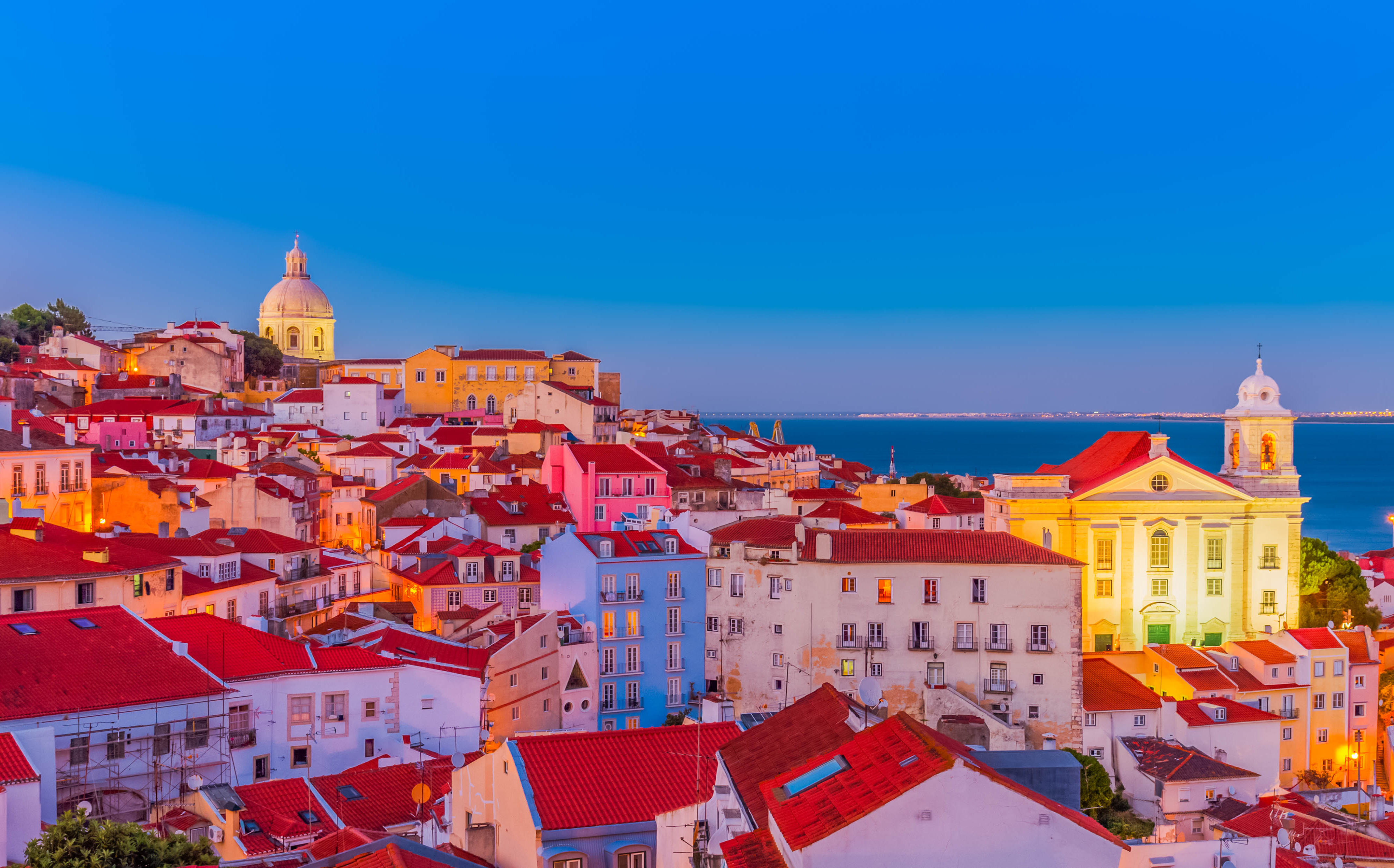 Romantisk panoramaudsigt over Lissabons ikoniske Alfama-kvarter ved blå time med farverige bygninger og havet i baggrunden