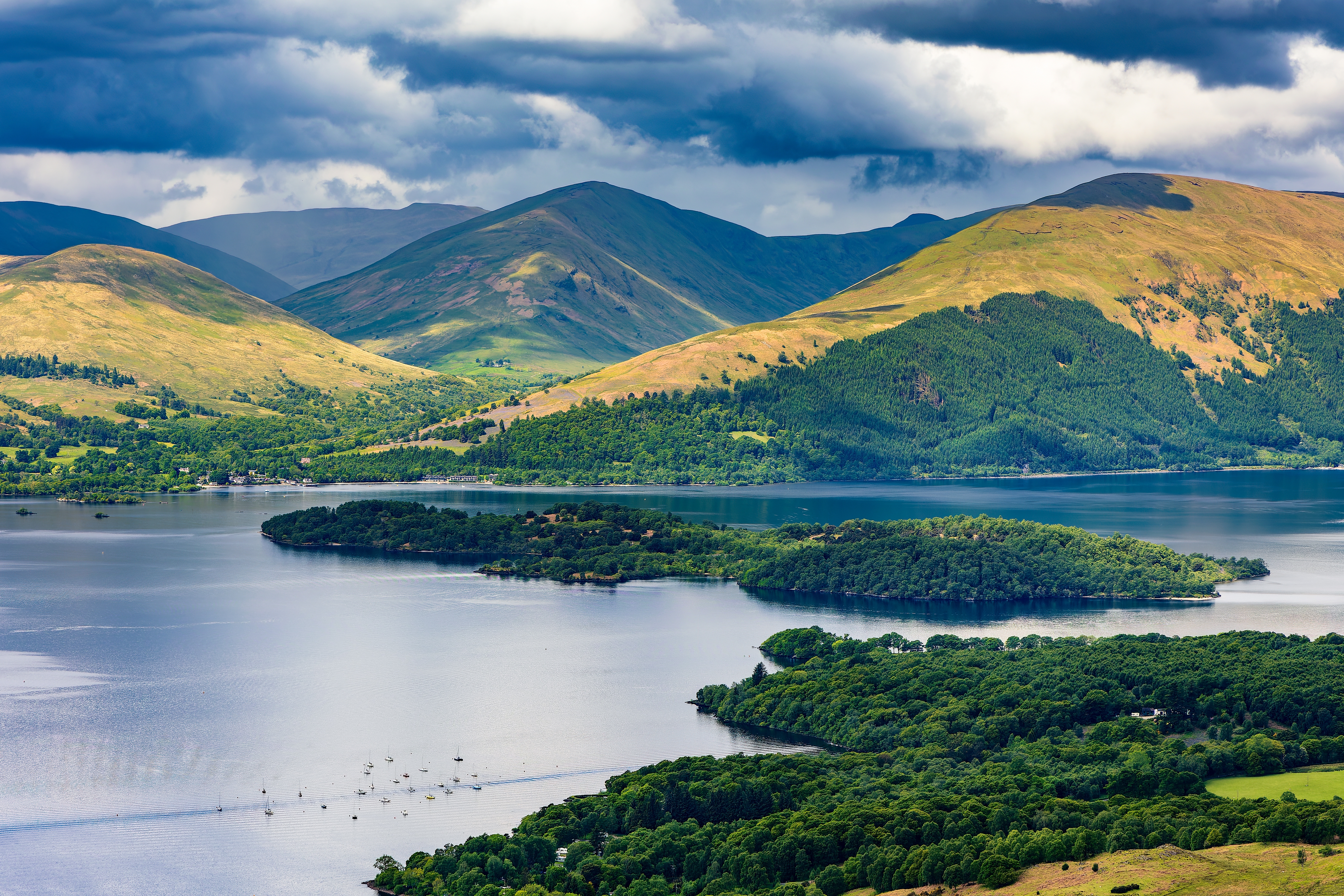 Udsigt over den smukke sø Loch Lomond i Skotland med grønne øer og høje bjerge under dramatisk himmel