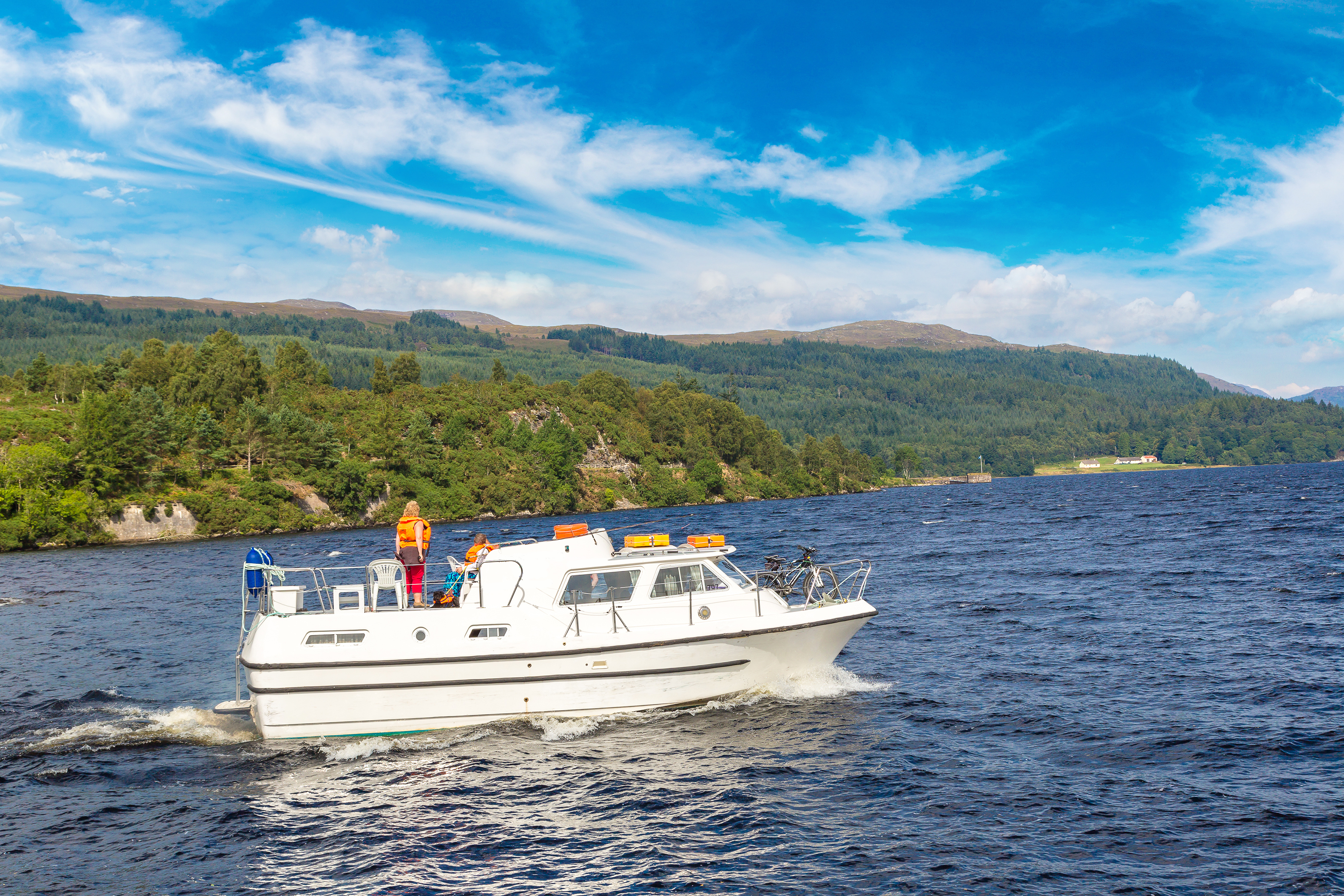 Hvid lystbåd sejler på Loch Ness i Skotlands højland med passagerer om bord og smukt naturlandskab i baggrunden