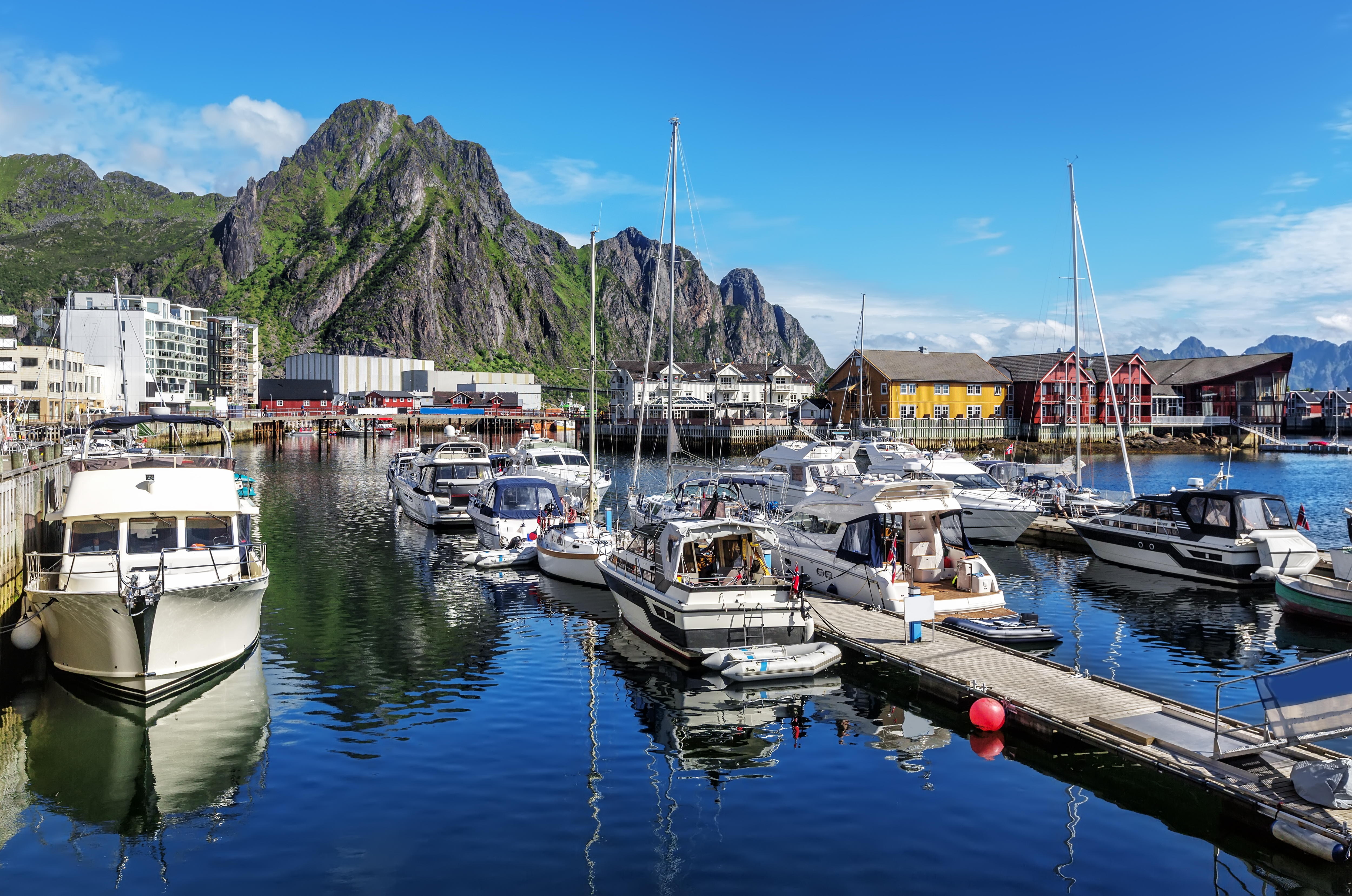 Smukt sommerbillede af Svolværs marina på Lofoten med både, farverige bygninger og de majestætiske norske bjerge under blå himmel