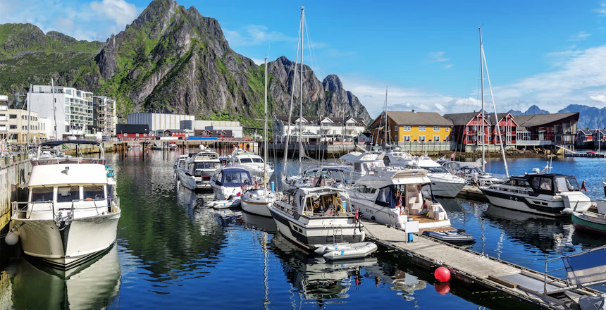 Smukt sommerbillede af Svolværs marina på Lofoten med både, farverige bygninger og de majestætiske norske bjerge under blå himmel