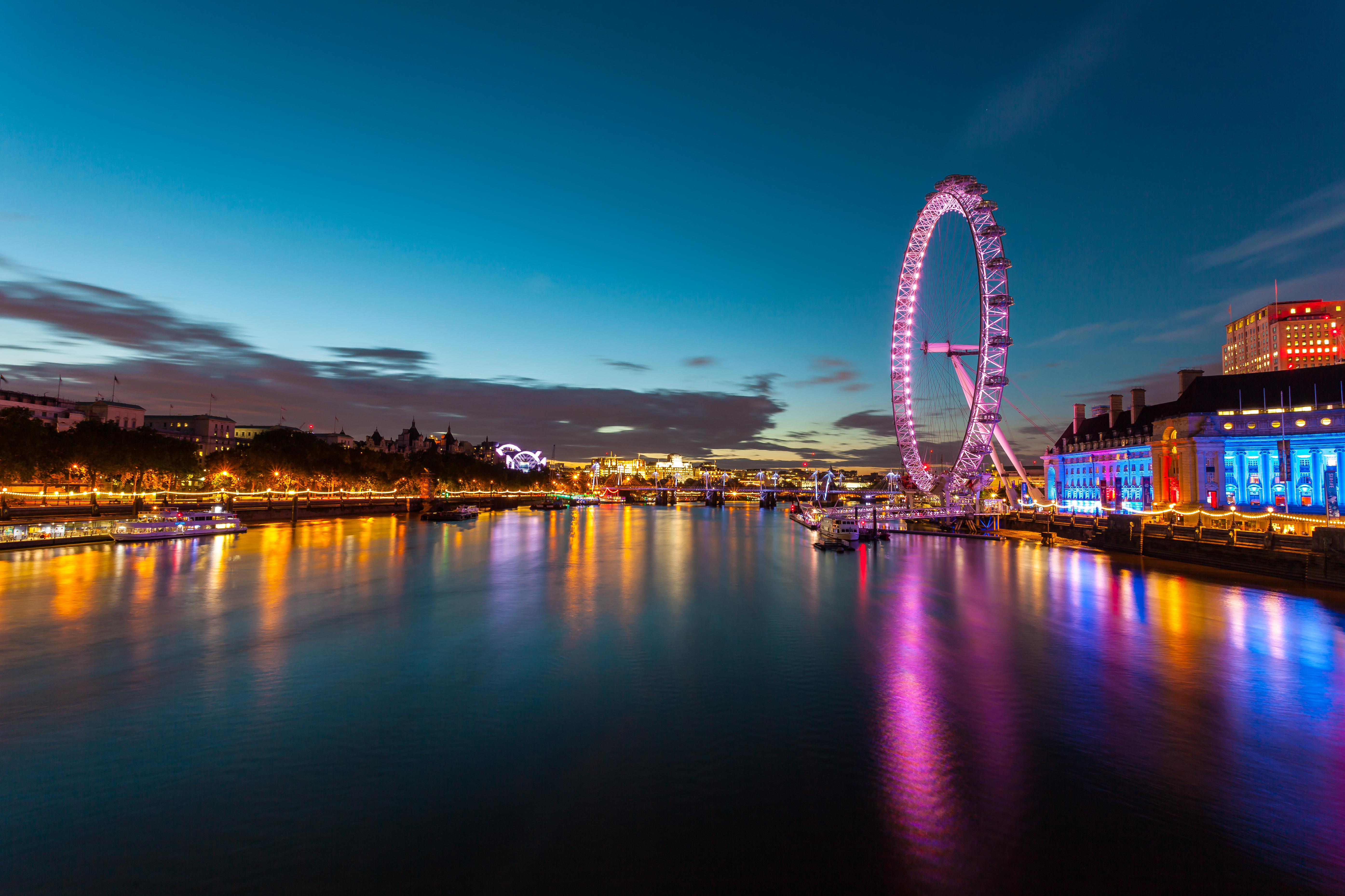 Udsigt over London Eye og Themsen om aftenen med farvestrålende lys og refleksioner i floden