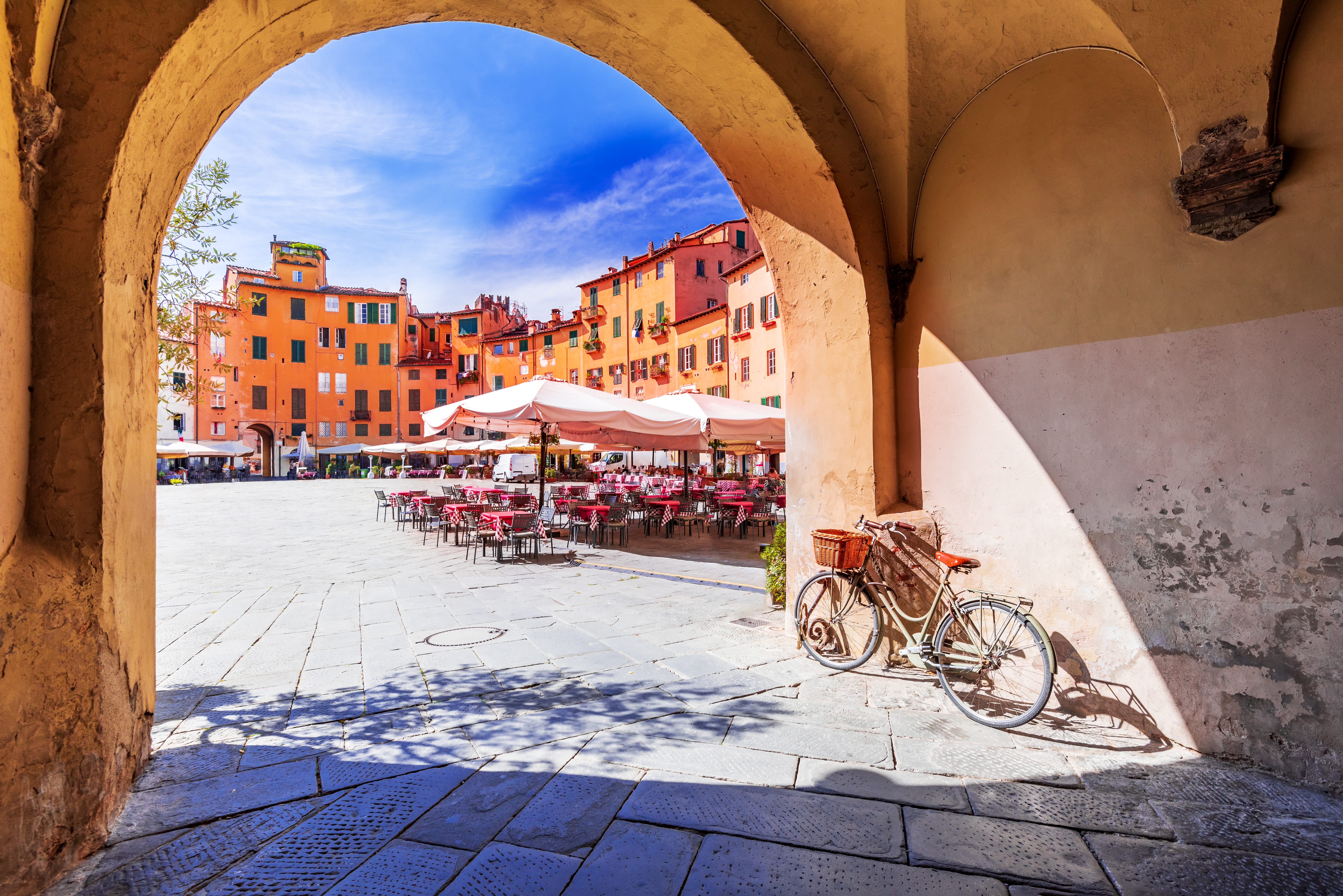 Det ovale Piazza dell'Anfiteatro i Lucca, Toscana, fotograferet gennem en historisk stenbue med en hvid cykel og charmerende café med røde borde